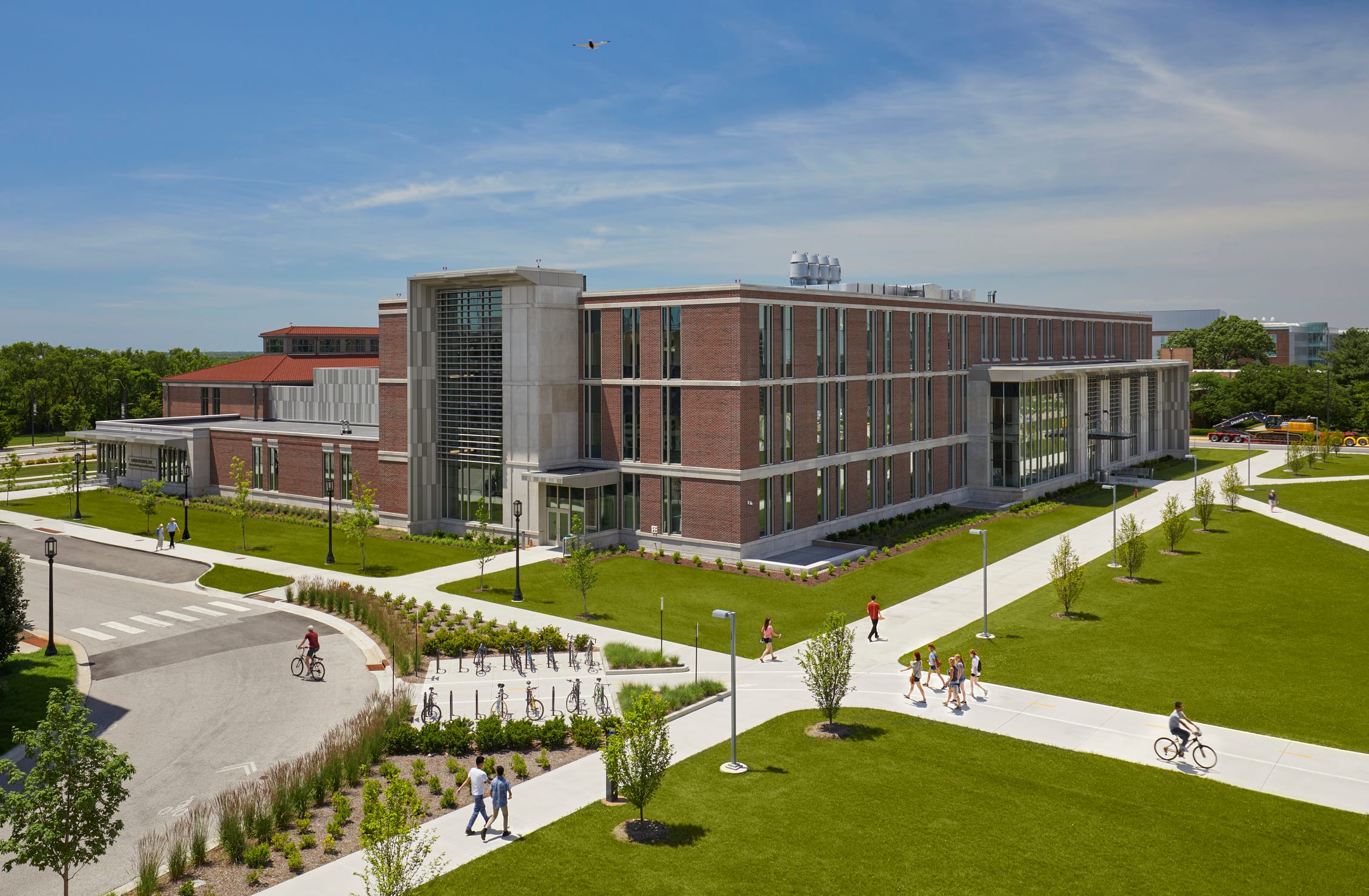 Aerial view of the Hobart and Russell Creighton Hall of Animal Sciences complex featuring a modern brick and concrete building with large glass entrances and window panels. The facility is surrounded by landscaped green spaces with newly planted trees, concrete walking paths, and a bicycle parking area. Students can be seen walking and cycling along the pathways under a clear blue sky. The architectural design combines traditional red brick with contemporary concrete elements and large windows, creating an inviting academic environment on campus.