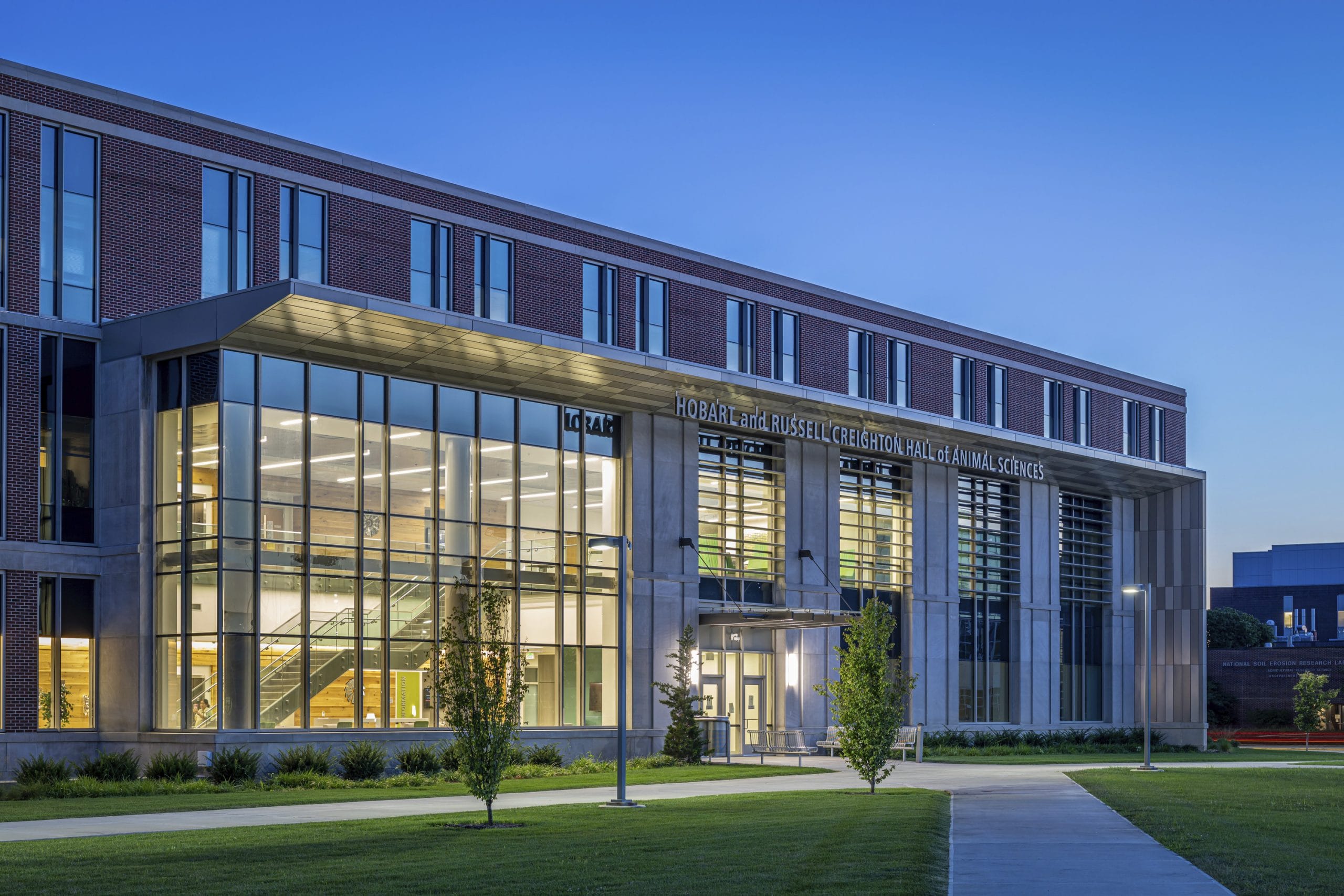 Exterior view of the Hobart and Russell Creighton Hall of Animal Sciences complex featuring a modern brick and concrete building with large glass entrances and window panels. The facility is surrounded by landscaped green spaces with newly planted trees, concrete walking paths, and a bicycle parking area.