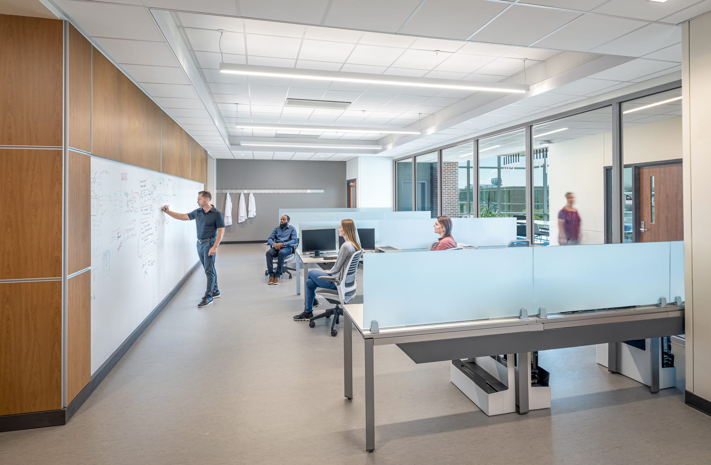 The image shows a modern educational or collaborative workspace. There are several desks with frosted glass panels situated in front of a white board. A man is writing something on the white board and appears to be teaching something to a group of three individuals situated at the desks. The space has a contemporary design with large windows along the right side behind the desks offering a view of the atrium and hallway. The overall impression is of a flexible learning environment or collaborative workspace.