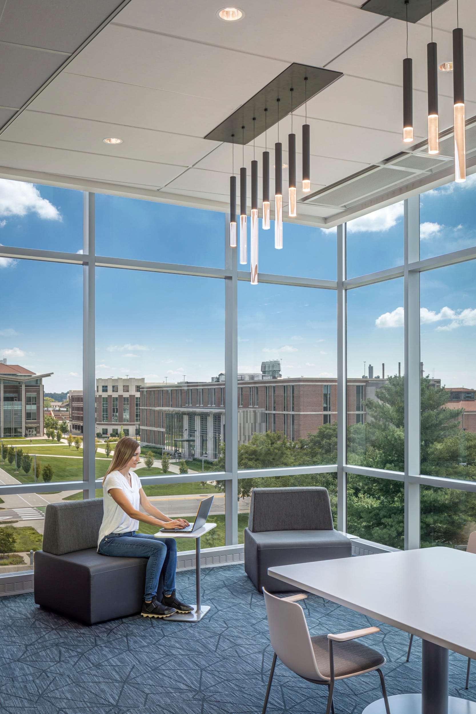 The image shows a section of a modern interior lobby area of the Martin C. Jischke Hall. The space has only floor-to-ceiling glass walls, allowing abundant natural light to flood the interior. The windows offer a stunning view of a well-maintained campus with impressive brick architecture. The lobby has a contemporary aesthetic with patterned blue carpeting. A woman is sitting in a modern grey chair working on a computer.