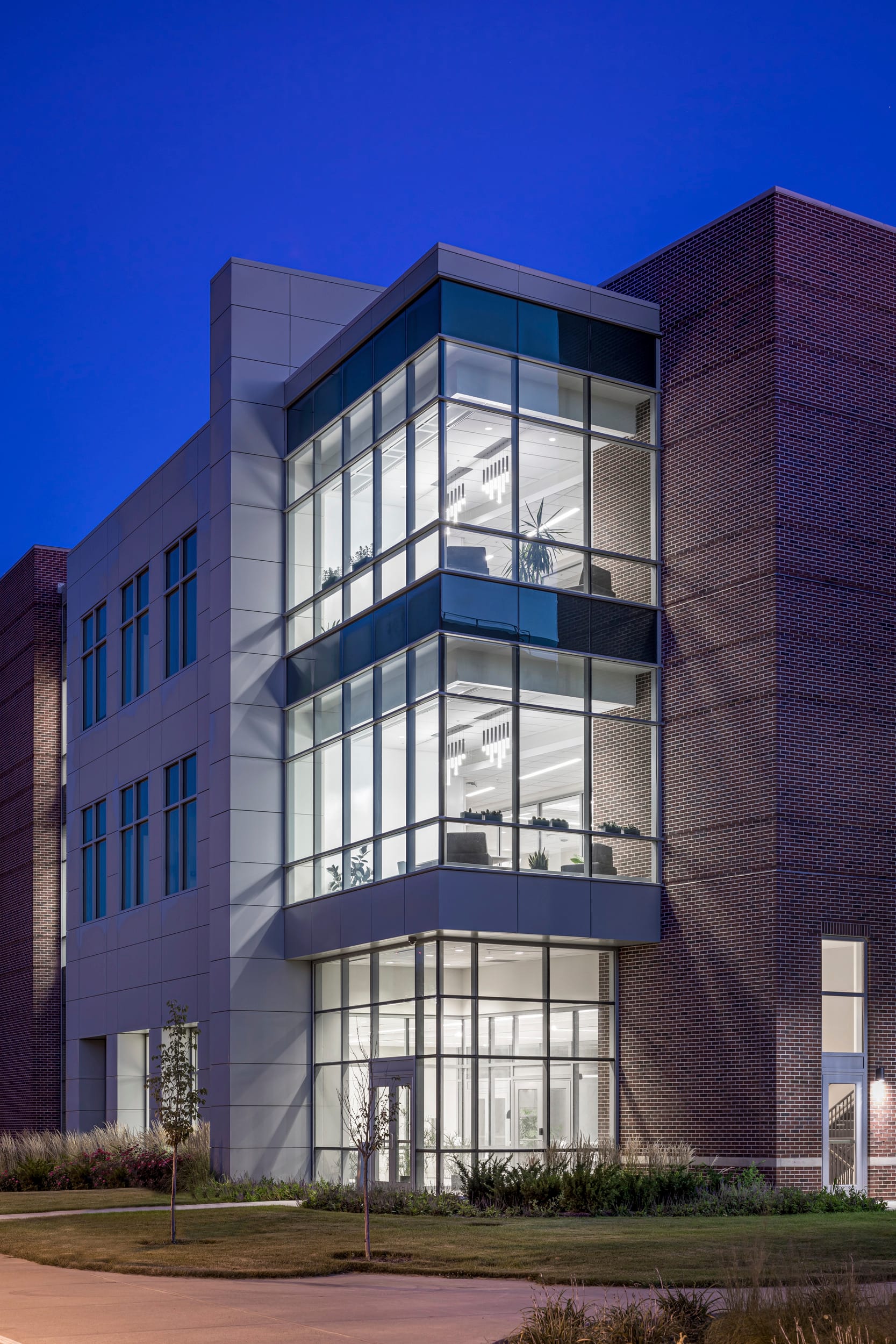 The image shows a modern three-story building photographed at dusk or early evening against a deep blue sky. The structure features a contemporary architectural design with a striking corner element made of floor-to-ceiling glass windows that project outward from the main structure. This glass corner provides a showcase view into the illuminated interior spaces on each floor. The building combines different exterior finishes, with red brick on the right side and light-colored panels or concrete on the left façade. The interior lights are on, revealing office or laboratory spaces with equipment visible through the large windows. The ground level appears to have an entrance area with glass doors. The landscaping around the building is minimal but includes some newly planted small trees or shrubs and ornamental grasses. A walkway or driveway is visible in front of the building. The overall impression is of a modern academic research facility, likely part of a larger campus complex based on the architectural style which appears consistent with the buildings shown in previous images.