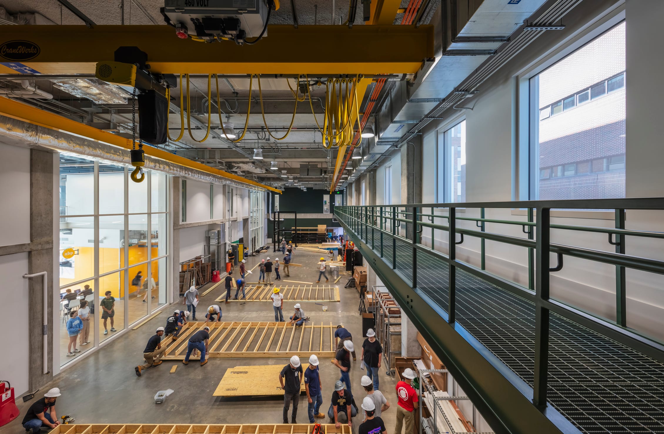 This image shows a construction laboratory inside Dudley Hall and Lambertus Hall at Purdue University. The high-ceilinged workshop space features yellow overhead cranes with hoists running along ceiling tracks, designed for lifting heavy materials. The main floor shows students wearing white hard hats working collaboratively on large wooden framing projects spread across the concrete floor. The perspective is taken from an elevated walkway with dark green railings that overlooks the workspace. Large windows along both walls allow natural light into the facility, with the familiar yellow design elements visible through glass walls on the left side. This appears to be a hands-on learning environment for construction or civil engineering students, providing practical experience with building techniques and materials. The industrial space is equipped with professional-grade equipment while maintaining visual consistency with the building's overall design aesthetic.