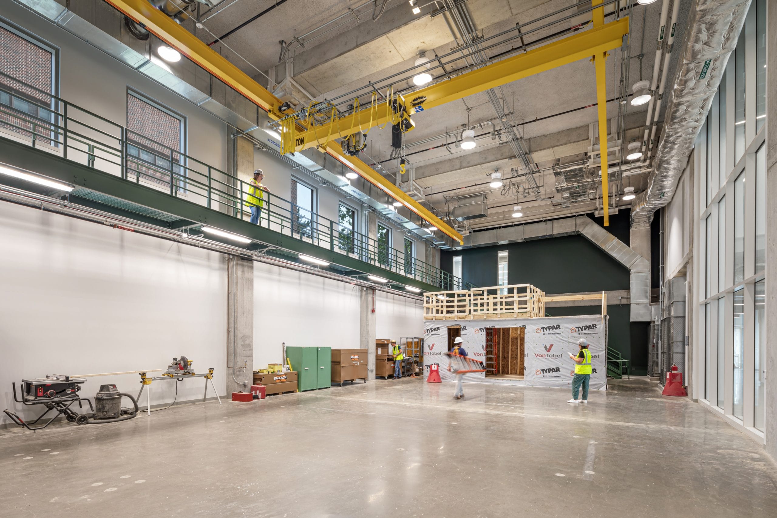 This image shows a construction laboratory inside Dudley Hall and Lambertus Hall at Purdue University. The high-ceilinged workshop space features yellow overhead cranes with hoists running along ceiling tracks, designed for lifting heavy materials. The main floor shows three students, one carrying a ladder, wearing white hard hats working collaboratively on large wooden framed structure. Above them is an elevated walkway with dark green railings that overlooks the workspace. This appears to be a hands-on learning environment for construction or civil engineering students, providing practical experience with building techniques and materials. The industrial space is equipped with professional-grade equipment while maintaining visual consistency with the building's overall design aesthetic.