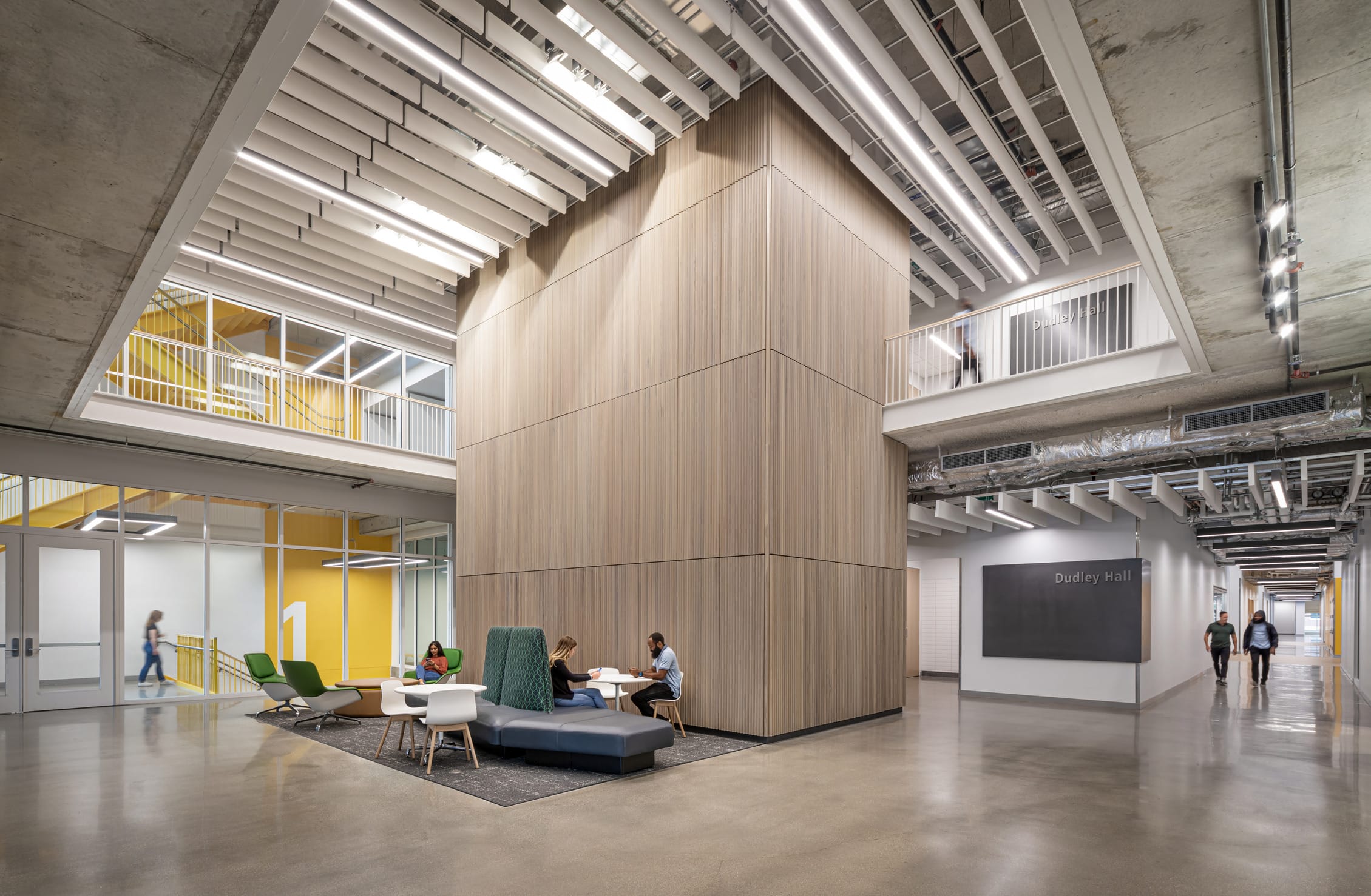 This image shows another interior space within Dudley Hall at Purdue University. The architecture features a central wooden-clad structure rising through multiple floors, with a contemporary open layout around it. A small collaboration area with colorful furniture (including green chairs, a blue bench, and a white table) is positioned in front of the wooden structure, where several students are working together. The distinctive yellow staircase seen in the previous image is visible through glass walls on the left. The space has polished concrete floors, a minimalist white ceiling with integrated linear lighting, and exposed mechanical systems. A 