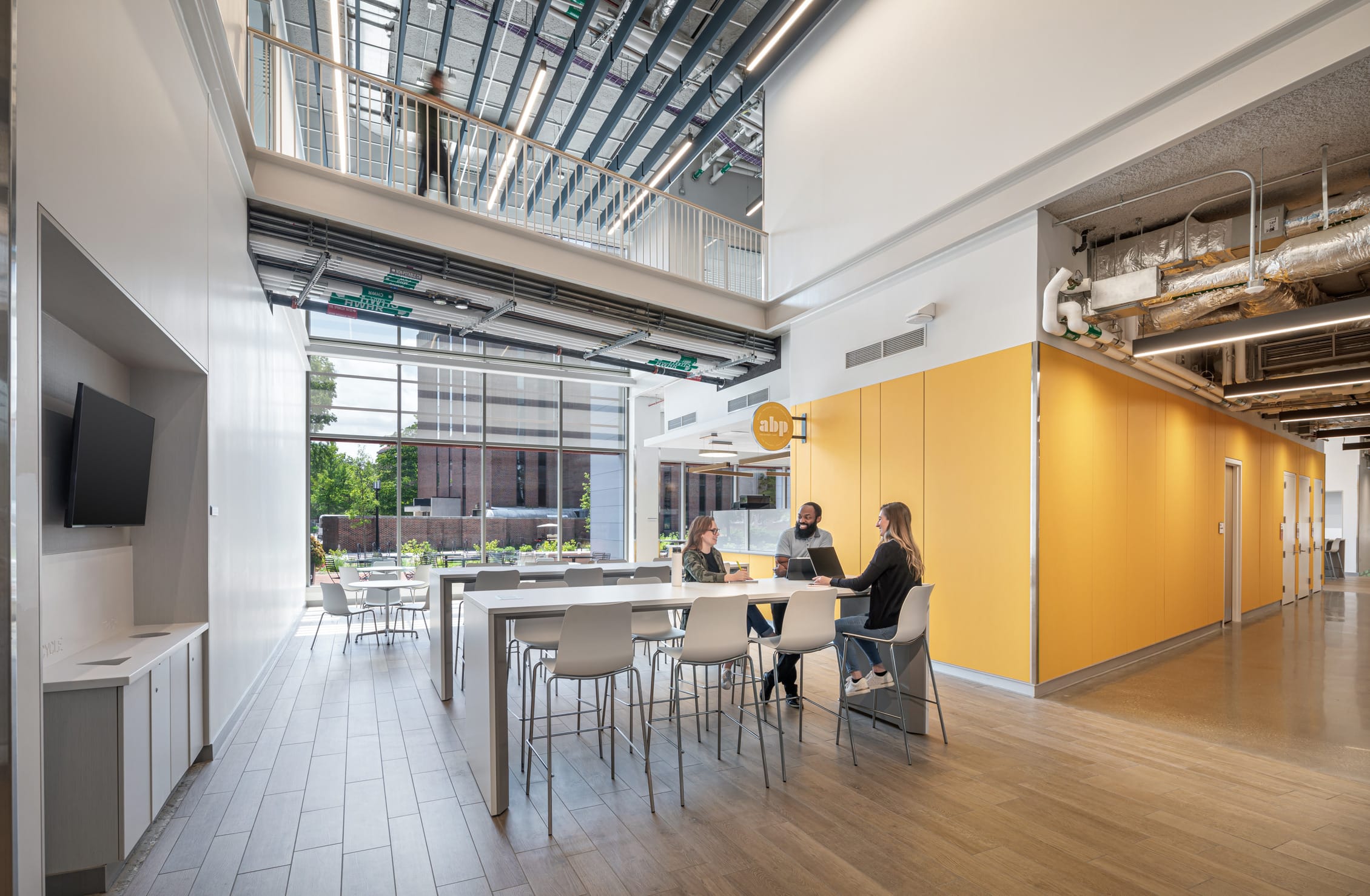 This image features a spacious, modern café with high ceilings, large windows, and yellow accents. Three people sit at a table, evoking a collaborative, bright atmosphere. The space has wood laminate floors and an industrial ceiling with exposed mechanical systems. Floor-to-ceiling windows in the background, beyond multiple sets of white café tables, allow natural light to flood into the space.