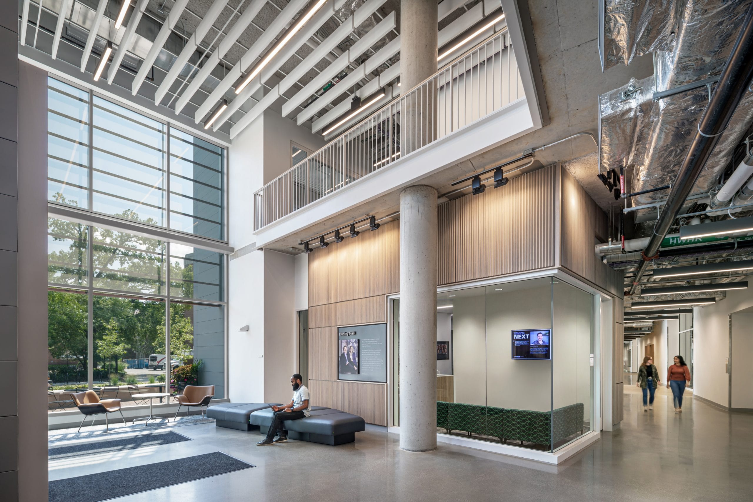 This image shows an interior space within Dudley Hall at Purdue University. The architecture features a central wooden-clad structure with a large concrete pillar ascending multiple levels. A small collaboration area with brown chairs and a large grey ottoman are positioned in front of the structure. Large floor-to-ceiling windows to the left of the structure allow natural light to flood into the building. The space has polished concrete floors, a minimalist white ceiling with integrated linear lighting, and exposed mechanical systems.