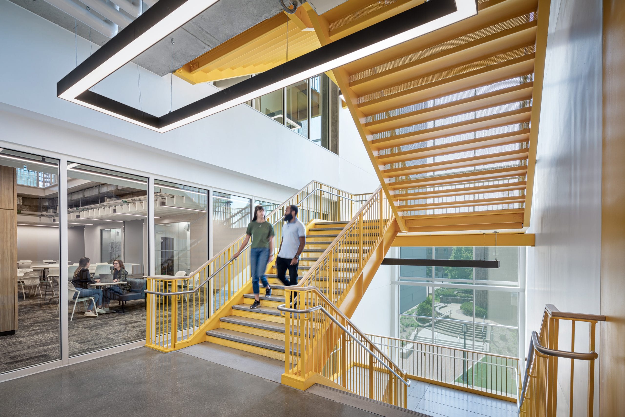 This image features a striking bright yellow staircase in a corridor with white walls and large windows running the full height of the multi-level stairwell. The expansive windows flood the space with natural light. On the left side of the stairwell, a modern collaborative area is visible through a full glass wall, furnished with sleek white chairs and tables. Two people are seen descending the stairs, engaged in conversation.