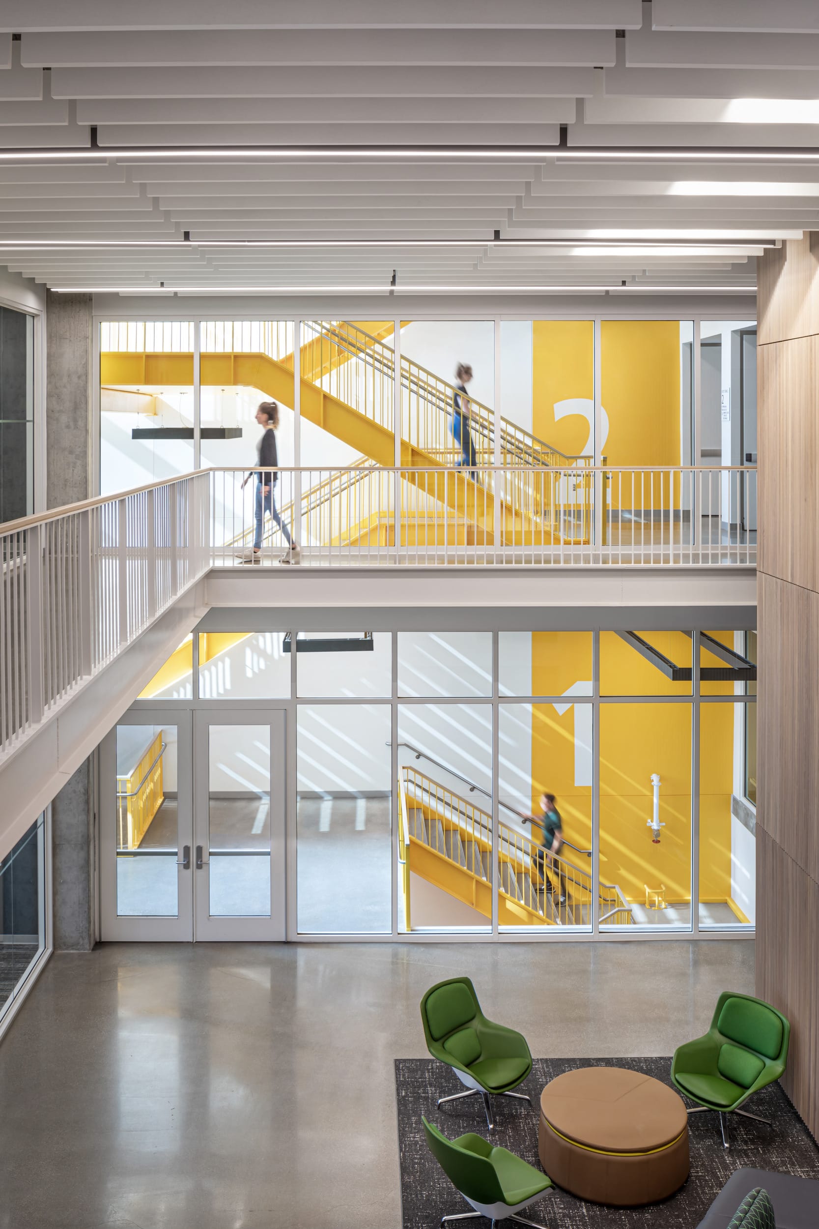 This image shows an interior view of Dudley Hall at Purdue University. The modern architecture features a striking yellow staircase visible through large glass walls that spans multiple floors. The view is taken from a lobby area with polished concrete floors where three bright green lounge chairs and a round tan ottoman create a small seating area. The space has a contemporary aesthetic with clean lines, white railings along an upper walkway, and a rhythmic white ceiling with integrated lighting. Two people can be seen in motion on the yellow staircase, slightly blurred as they move. The design creates visual interest through the contrast of the bright yellow architectural element against the otherwise neutral palette of white, gray, and light wood finishes on the walls.