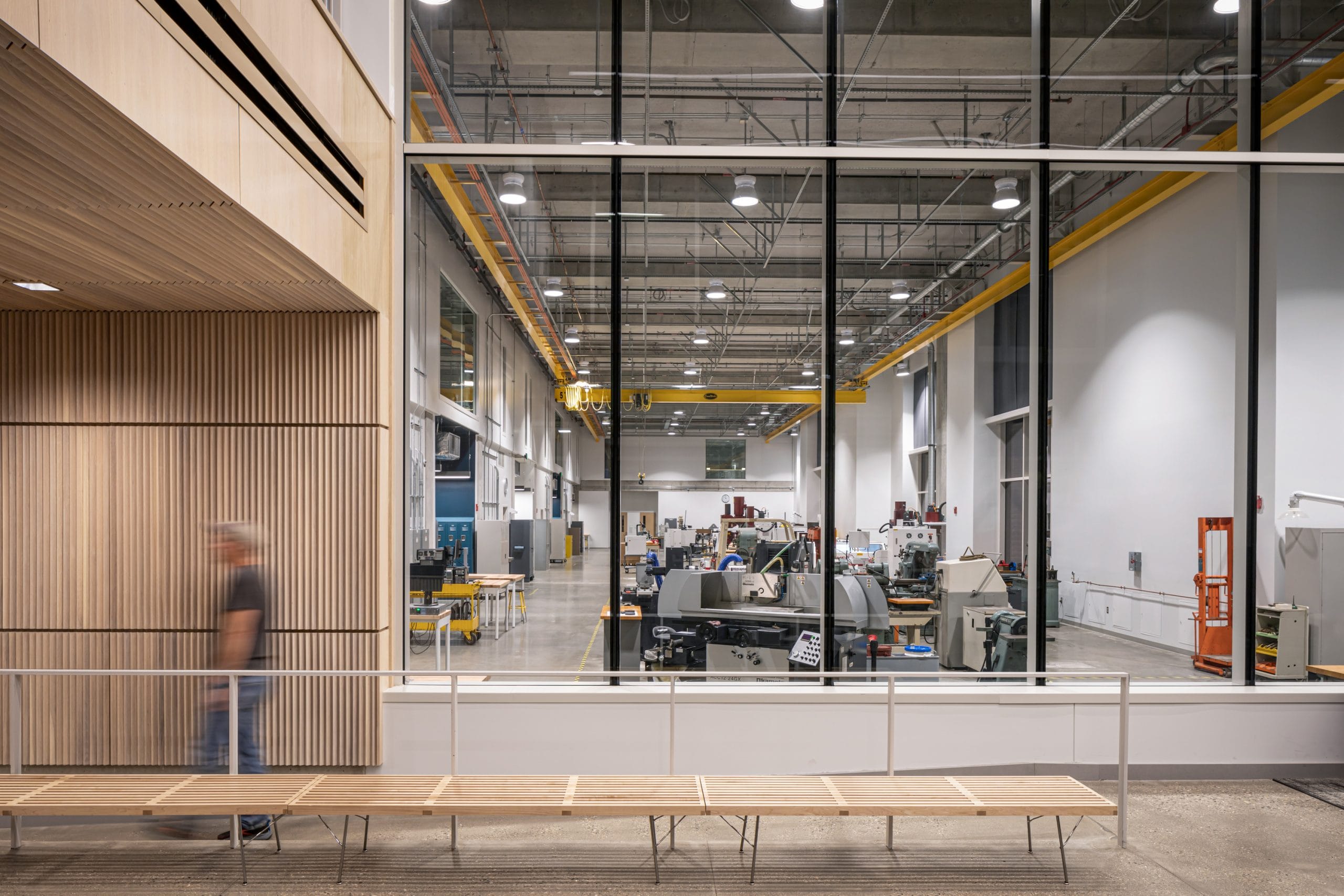 A modern industrial workspace viewed through large floor-to-ceiling windows. Inside the high-ceilinged manufacturing lab, various machinery and workstations are neatly arranged. Overhead, exposed ductwork and bright lights contribute to a functional, clean aesthetic. A yellow crane rail runs along the ceiling. In the foreground, a person walks past a minimalist wooden bench set against a wall of vertical wooden slats, highlighting the blend of industrial and contemporary design.