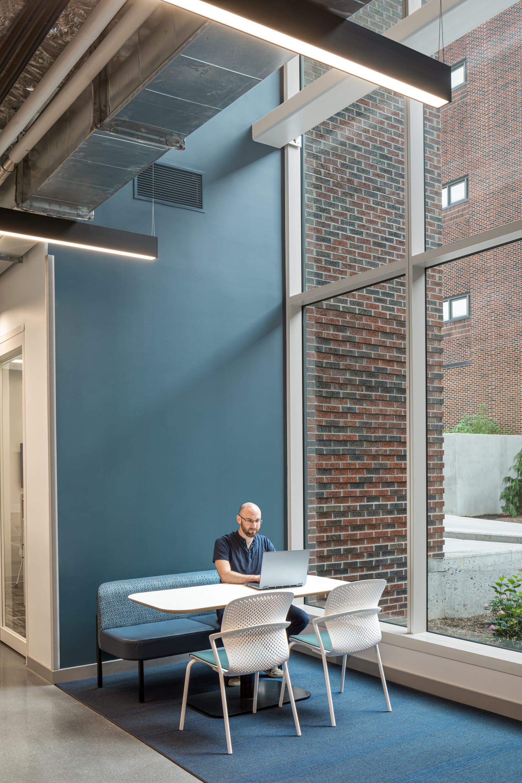 A modern student lounge area featuring a mix of blue tones, with a small couch and two chairs with a shared workstation arranged on blue carpet. Above, acoustic ceiling baffles and exposed piping add an industrial feel. A flat screen is mounted on a dark blue accent wall. There are floor-to-ceiling windows next to the work area.