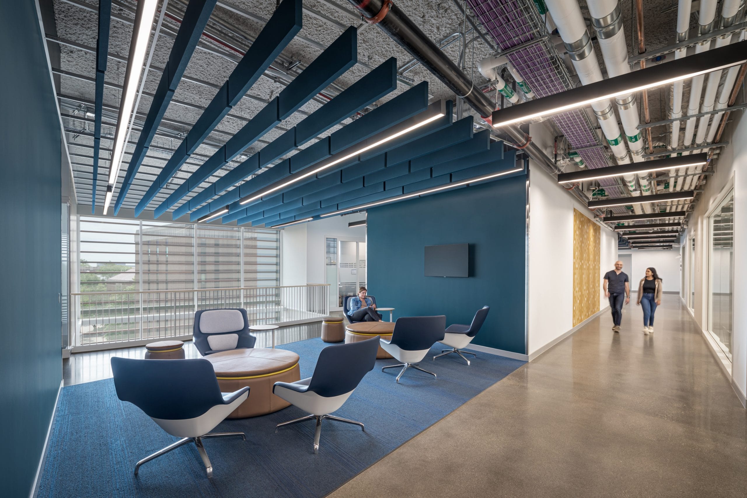 A modern student lounge area featuring a mix of blue tones, with stylish chairs arranged around round ottomans on a blue carpet. Above, acoustic ceiling baffles and exposed piping add an industrial feel. A flat screen is mounted on a dark blue accent wall. Large windows allow natural light to pour in. On the right, a polished concrete hallway leads past a decorative wooden wall, where two people are walking together.