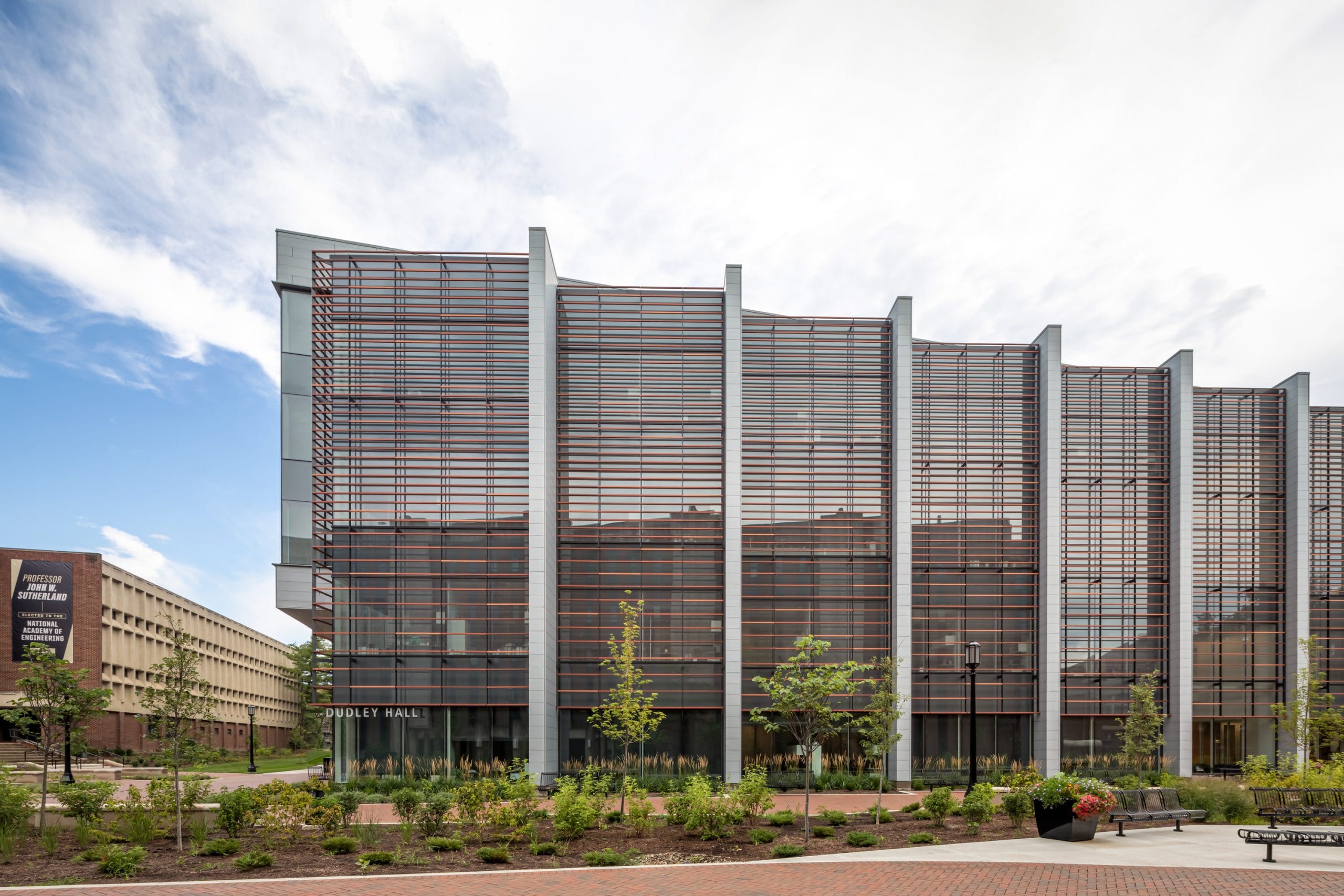 This image shows the exterior of Dudley and Lambertus Hall at Purdue University. The modern building features an impressive façade with extensive glass curtain walls, vertical metal sunshades, and a contemporary architectural design that combines brick elements with sleek contemporary materials. The entrance, marked 