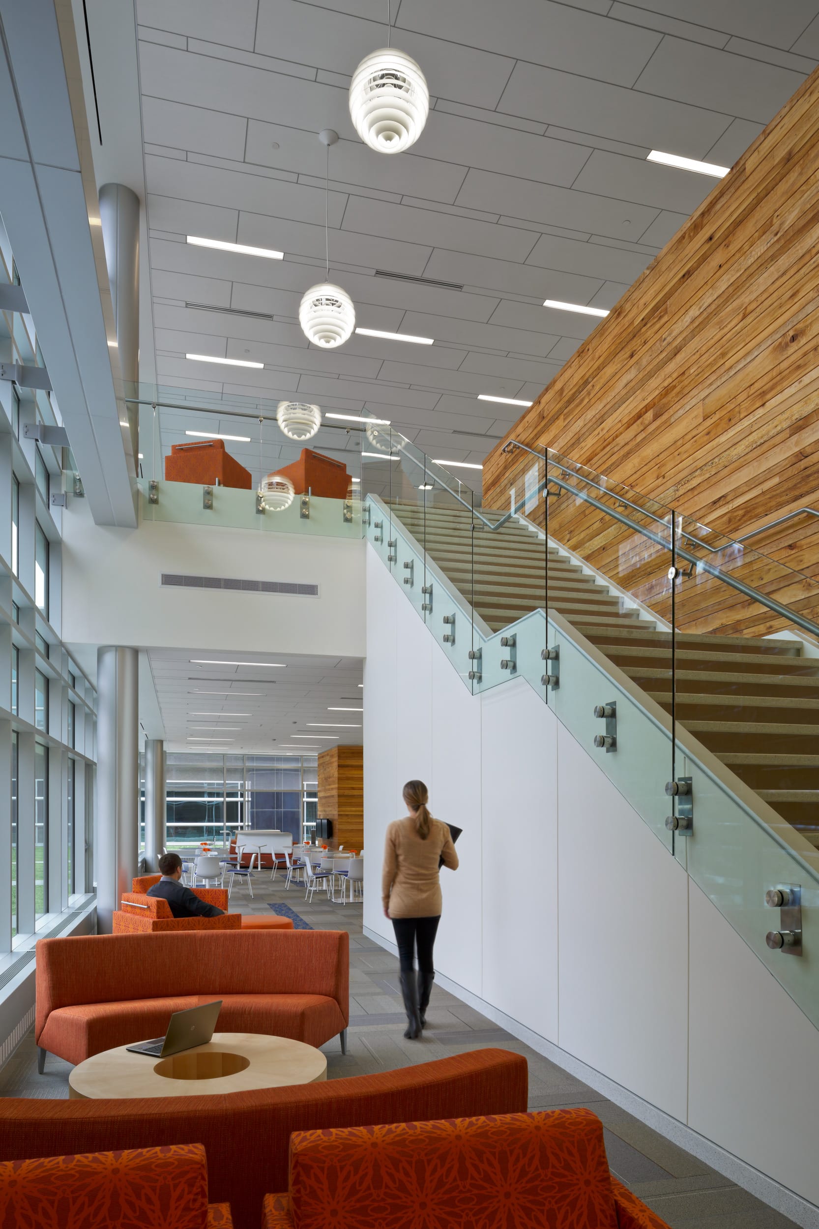 This image shows the interior of a modern institutional building featuring a stylish open-concept design. In the foreground are bright orange lounge chairs and a seating area with a round coffee table. A person in light-colored clothing is walking toward a modern staircase with glass railings and wooden treads that ascends along a white wall on the right side. The space has high ceilings with contemporary spherical pendant lights hanging from above. The left side features large windows letting in natural light. In the background, more orange furniture is visible in what appears to be a communal area. The combination of wood, glass, and white surfaces creates a clean, contemporary aesthetic that's characteristic of modern research facilities designed to promote collaboration and innovation.