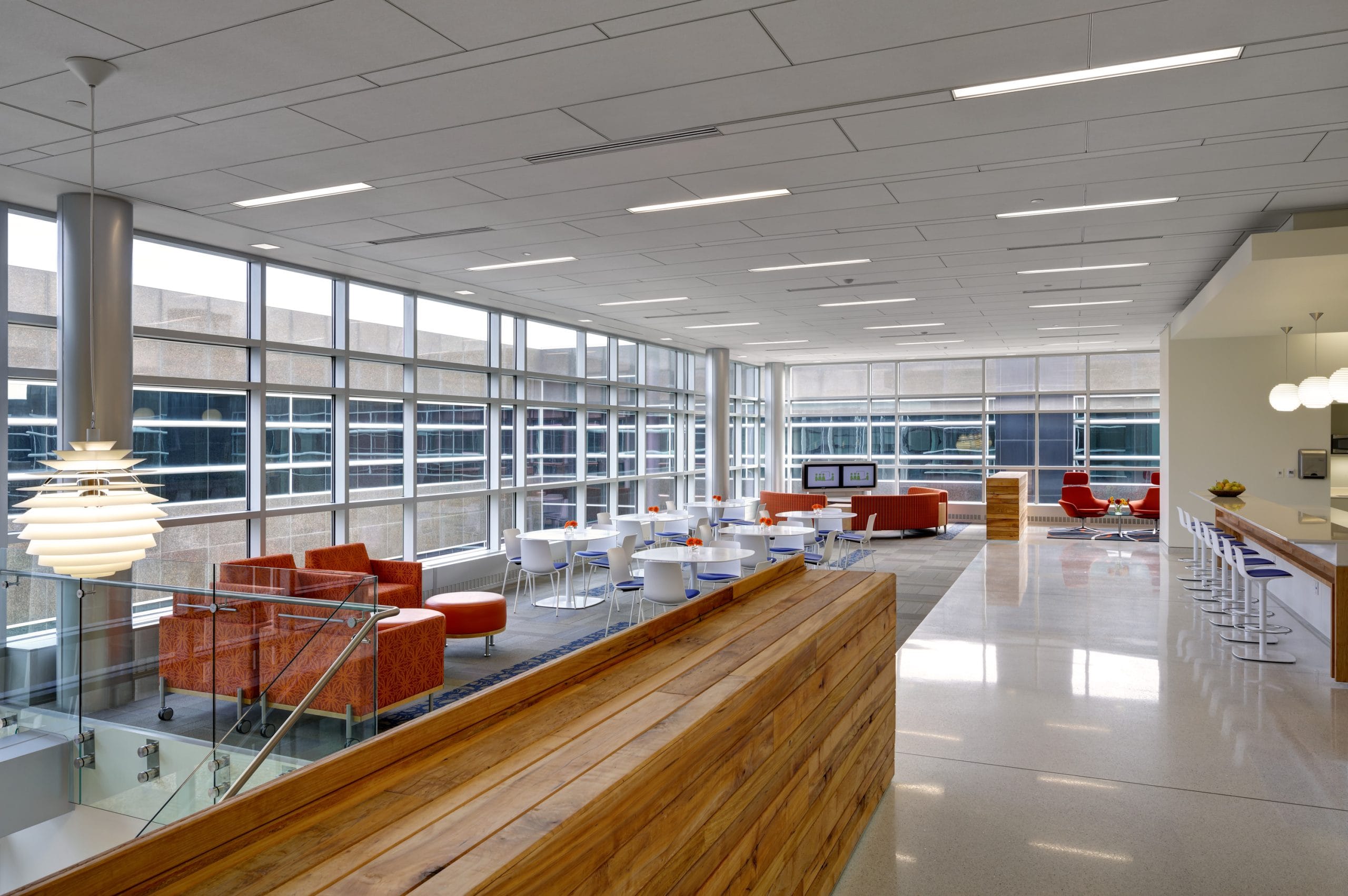 This image shows the second floor interior of a modern institutional building featuring a stylish open-concept design. In the foreground is a modern wood divider separating the descending stairs and the hallway. There are also bright orange lounge chairs and a red ottoman. In the background, more orange furniture as well as white and blue furniture are visible in a communal area. Floor-to-ceiling windows throughout the entire space allow light to flood in. This contemporary aesthetic is characteristic of modern research facilities designed to promote collaboration and innovation.