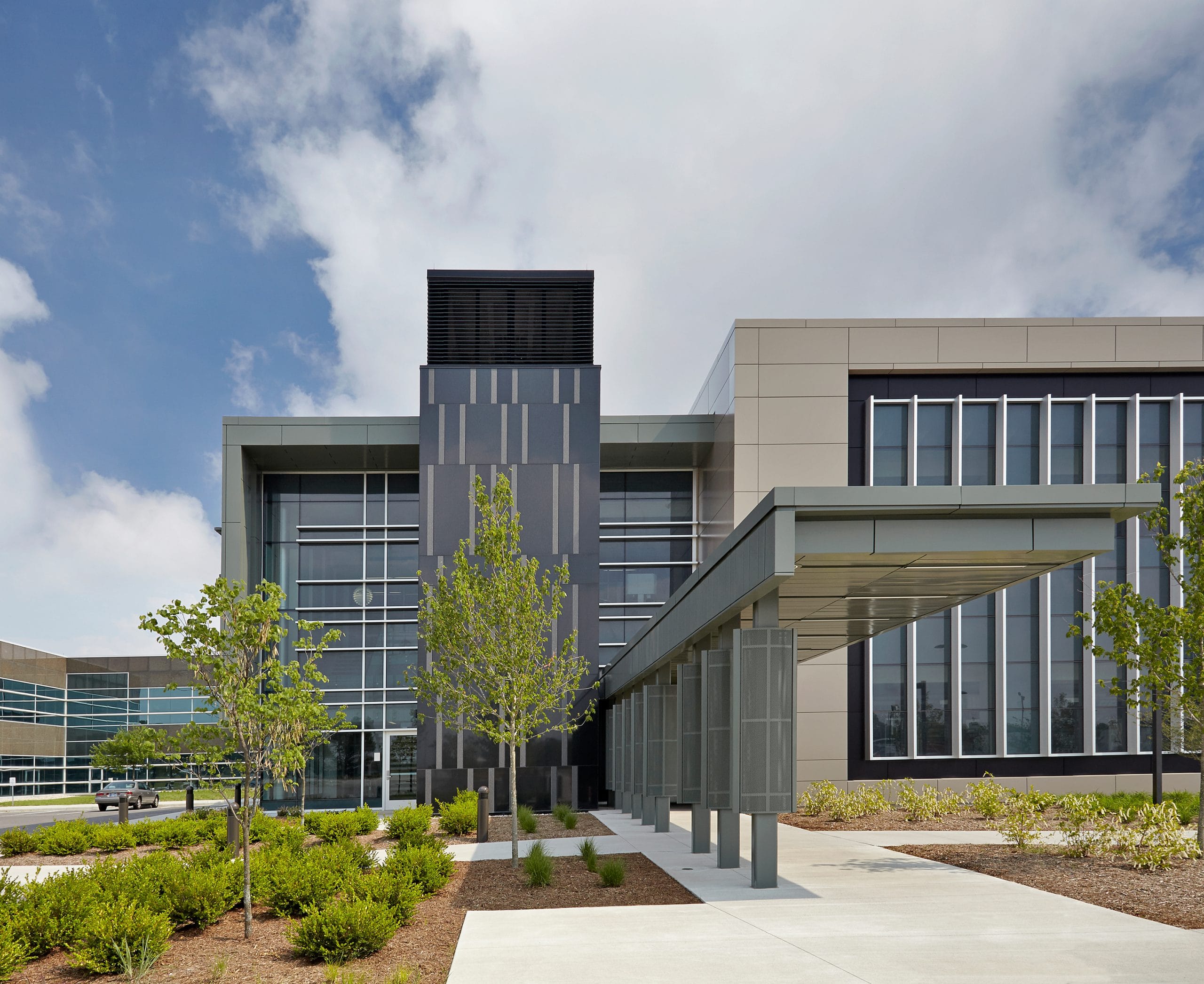 A research building with a sleek façade featuring a mix of glass, metal, and light-colored panels. The entrance is highlighted by a covered walkway with a metal canopy supported by perforated columns. The landscaping includes neatly arranged trees, shrubs, and mulch beds. Large windows reflect the partly cloudy sky, and the overall aesthetic is contemporary with a strong geometric design.