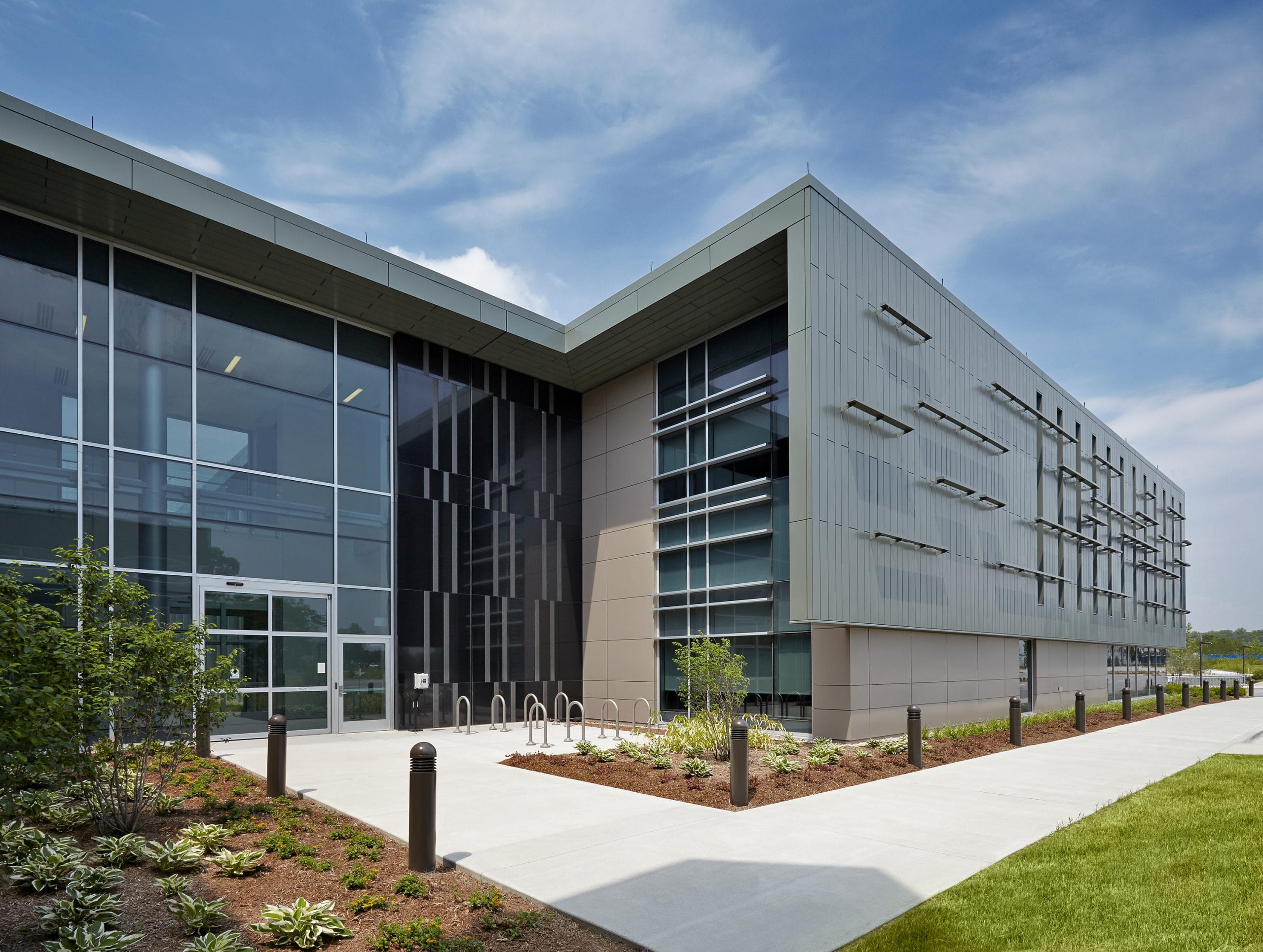 A research building with a sleek façade featuring a mix of glass, metal, and light-colored panels. The landscaping includes neatly arranged trees, shrubs, and mulch beds. Large windows reflect the partly cloudy sky, and the overall aesthetic is contemporary with a strong geometric design.