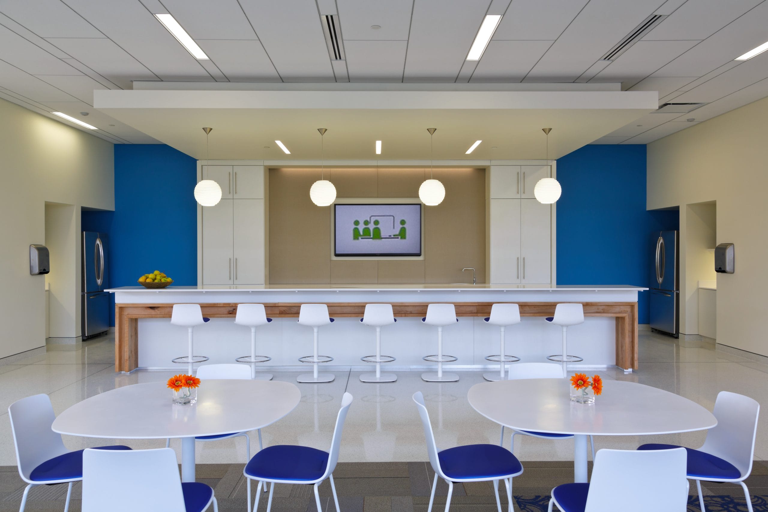 modern and well-lit break room or office cafeteria with a spacious, open layout. A long bar-style counter with white barstools lines the back wall, with pendant lights hanging above. The counter is backed by white cabinets, a television screen displaying a simple icon-based presentation, and blue accent walls. Two refrigerators are positioned in alcoves on either side of the counter. In the foreground, white tables with blue chairs are arranged neatly, with small orange flower vases adding a touch of color. The floor is made of a polished material, reflecting the bright ceiling lights, contributing to the sleek and professional ambiance.