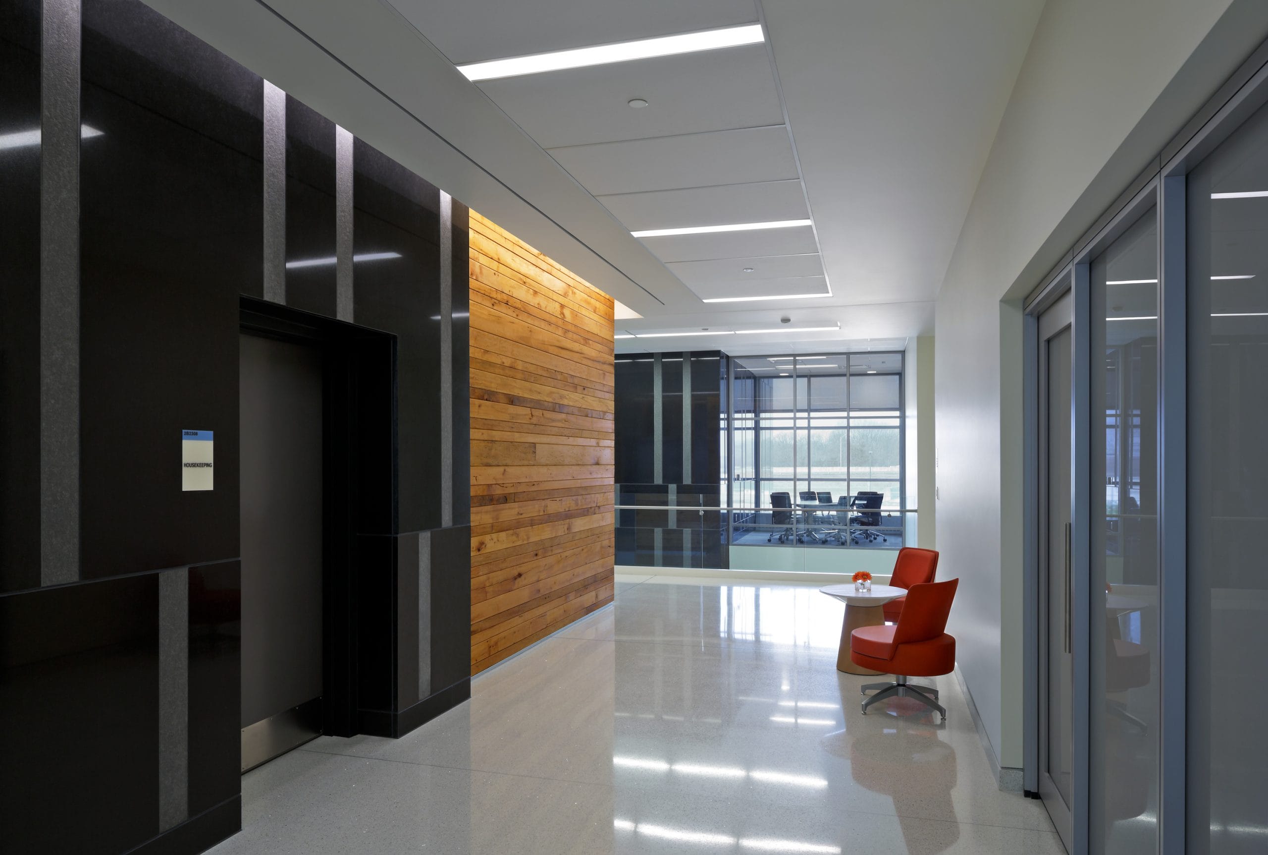 This image shows a modern, minimalist corridor or reception area in Building 314. The space features sleek design elements including a black wall panel on the left side, contrasted with a warm wooden accent wall made of horizontal planks. The floor is polished white, creating a reflective surface that enhances the brightness of the space. In the foreground to the right, there are two vibrant red chairs positioned next to a small round table, creating a small waiting area. Through glass partitions in the background, a conference room or meeting space with dark chairs is visible. The ceiling has recessed lighting fixtures that illuminate the hallway evenly. The overall aesthetic combines industrial elements with warm natural materials, characteristic of modern architectural design.
