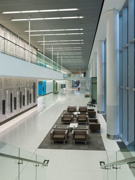A spacious hospital lobby with high ceilings, sleek modern design, and large floor-to-ceiling windows allowing natural light. The area features contemporary seating arranged on a dark rug, with white tile flooring and a glass railing leading to a staircase. The walls are decorated with textured panels, and overhead lighting lines the ceiling. A second-floor walkway with glass barriers is visible on the left. This image showcases BSA's expertise in interior architecture and design.
