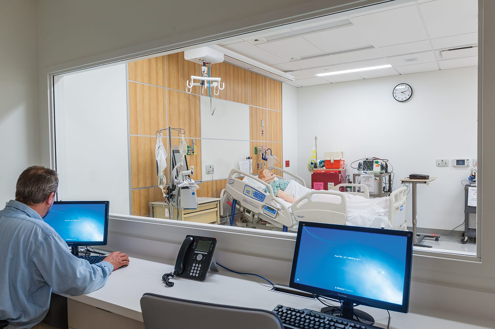 This image shows a medical simulation or training room with a partial view of a person at a monitoring station equipped with multiple computer screens and a desk phone, looking through a large observation window into a hospital room setup containing a medical mannequin in a hospital bed, surrounded by typical medical equipment including IV stands, monitors, and emergency equipment, with wood-paneled accent walls, ceiling-mounted equipment, and a wall clock, representing a clinical education environment likely used for healthcare professional training.
