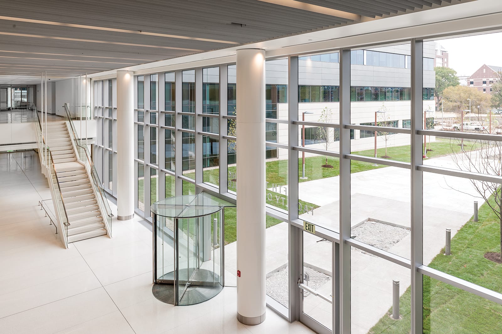 This image shows a bright, spacious corridor within the Fifth Third Faculty Office Building at Eskenazi, featuring floor-to-ceiling windows that provide views of a landscaped courtyard area outside with grass, walkways, and young trees; the interior hallway has white tiled floors, cylindrical support columns, a glass revolving door entrance, a staircase with glass railings on the left, and a modern slatted ceiling design, demonstrating the building's contemporary architectural style that prioritizes natural light and visual connection to outdoor spaces.