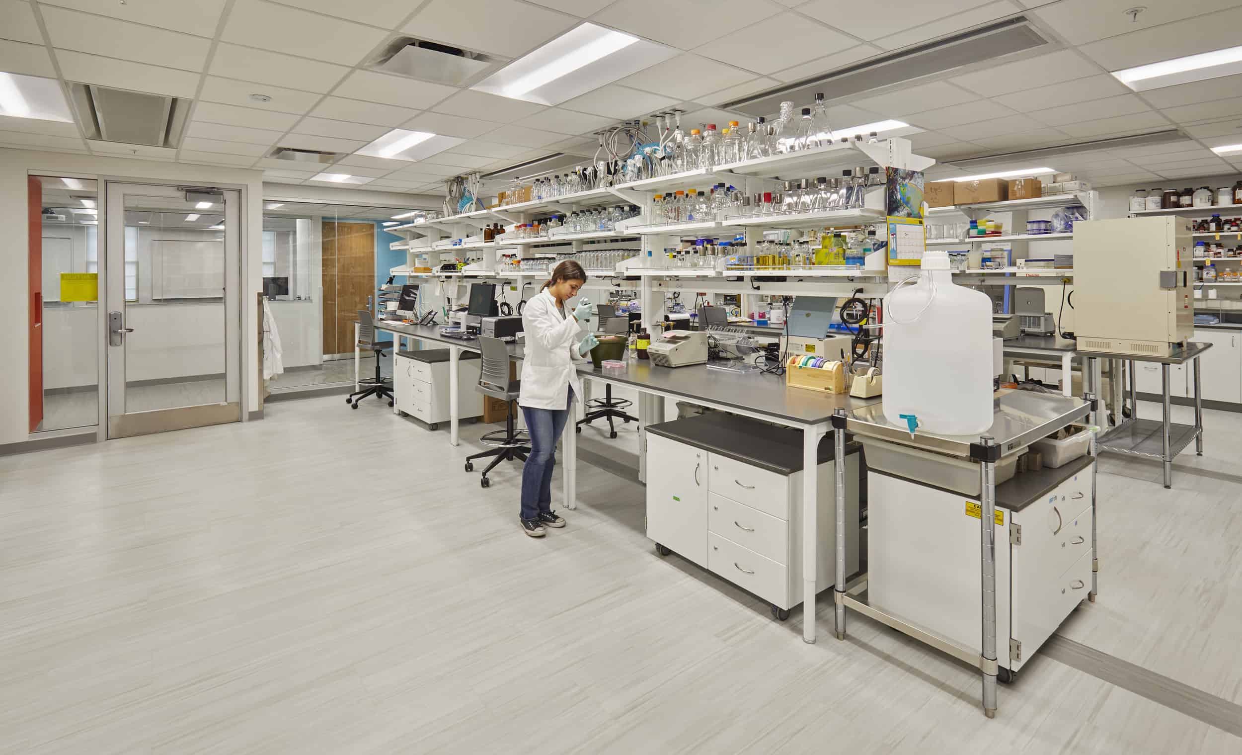 This image shows a well-equipped scientific laboratory space. A researcher in a white lab coat is working at one of the lab workstations in the center of the room. The lab features extensive shelving along the back wall filled with numerous glass bottles, containers, and laboratory glassware of various sizes. There are several laboratory workbenches with dark countertops and white cabinet storage underneath. A large water container can be seen in the foreground right. The lab has light-colored flooring, a drop ceiling with recessed lighting, and glass doors visible on the left that lead to another area. Various scientific instruments and equipment are placed throughout the workspace. The overall appearance is of a modern, functional wet lab designed for chemical or biological research.