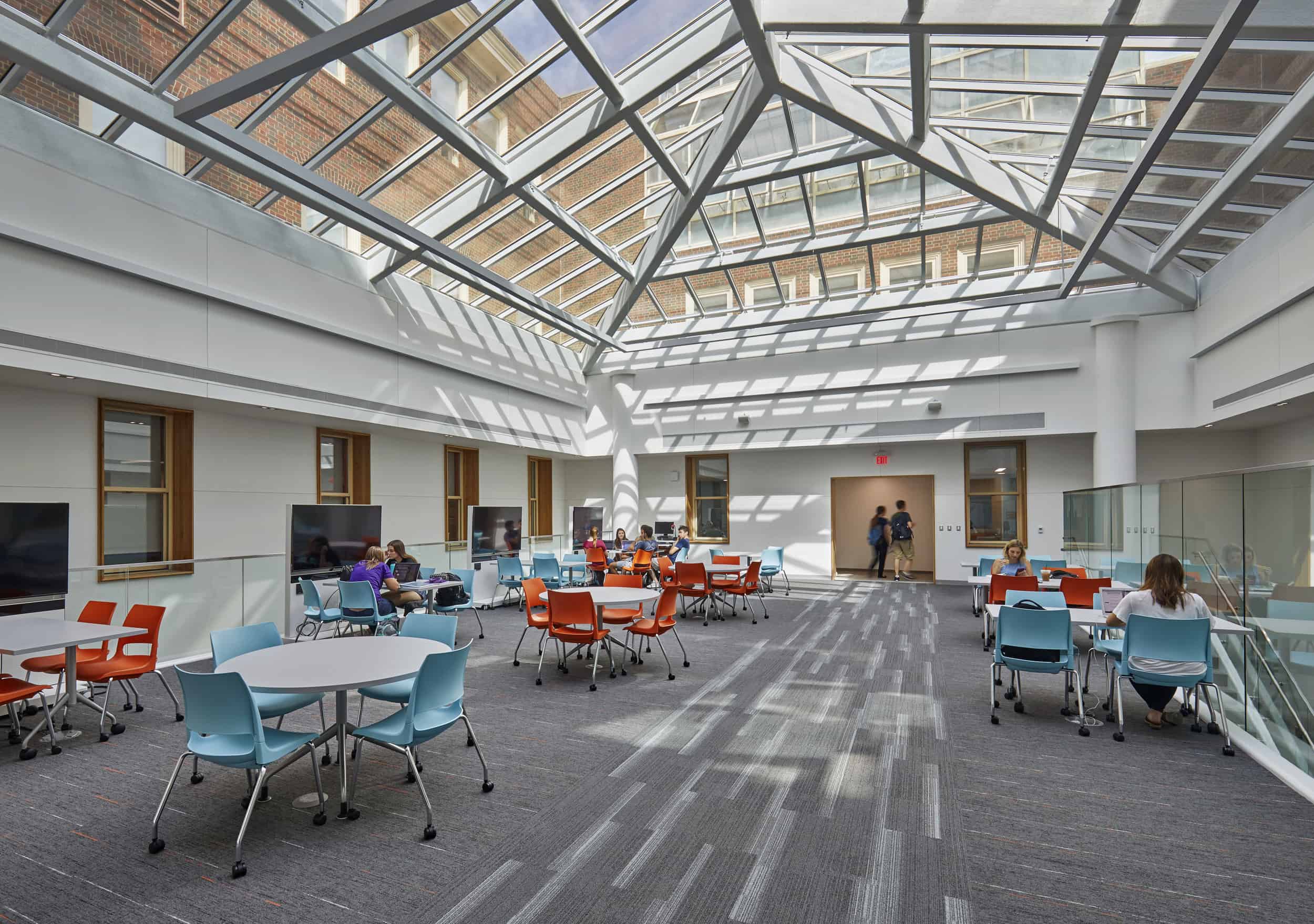 This image shows an open collaborative space in Hughes and Pearson Hall at Miami University. The area features a striking glass ceiling skylight with geometric metal framework that allows natural light to flood the space, creating distinctive shadow patterns on the floor. The room contains several round tables with brightly colored chairs in orange and light blue, arranged for group work or study. Several students can be seen sitting at these tables, working individually or in small groups. The space has white walls with wooden-framed windows, glass railings along what appears to be an upper level or mezzanine area, and a gray carpeted floor. The overall design creates a bright, airy atmosphere that appears conducive to collaboration and academic work.