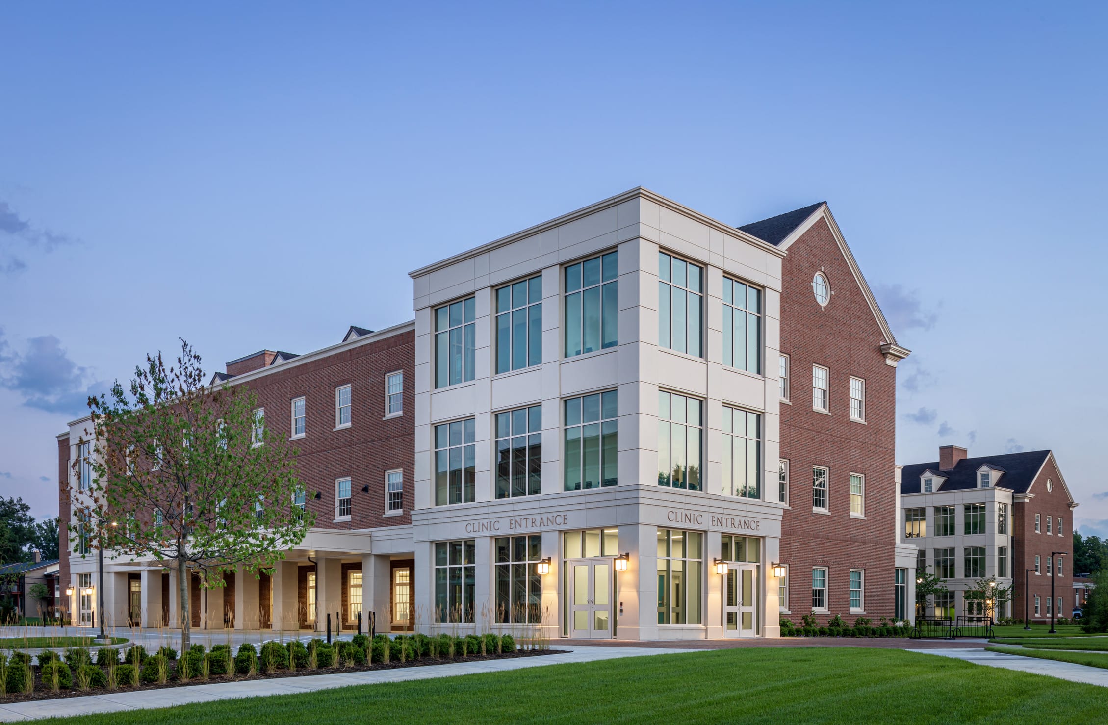 An evening exterior view of the Miami University Health Sciences Building clinic entrance, captured at dusk with the interior lights illuminating the glass façade. The architecture blends traditional collegiate elements with modern design, featuring red brick walls with a contemporary cream-colored corner entrance marked