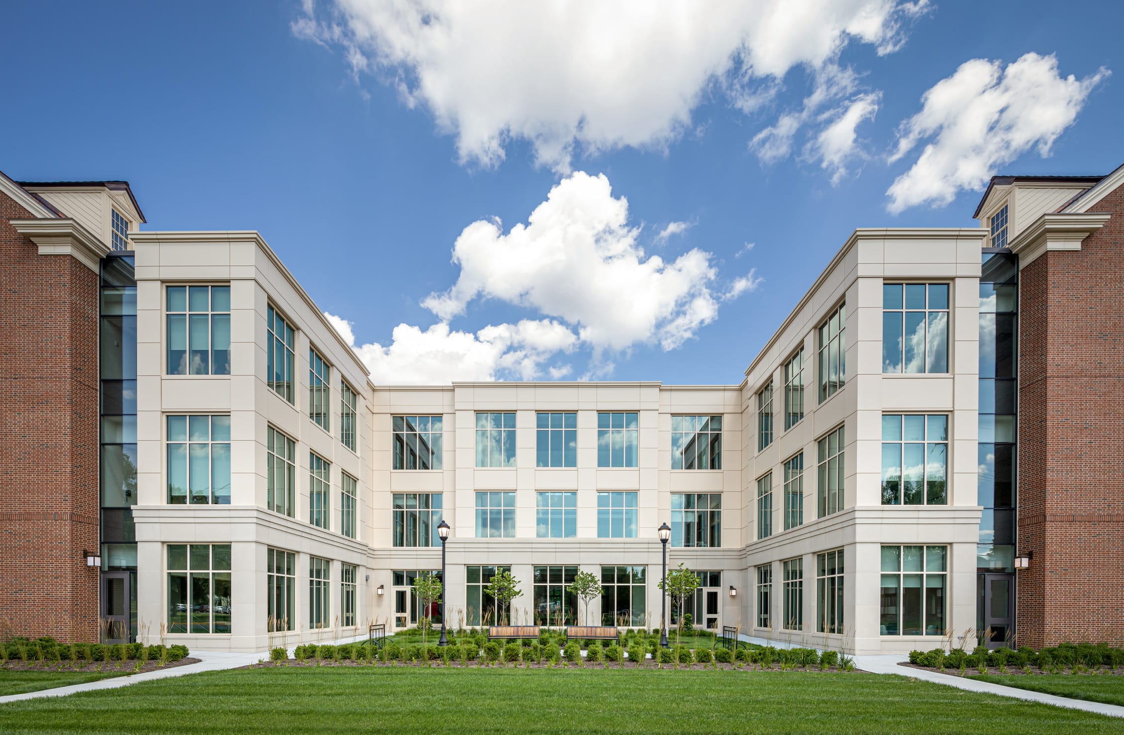 The exterior of Miami University's Health Sciences Building showcasing a modern U-shaped design with a combination of cream-colored stone and red brick facades. The four-story structure features abundant large windows that reflect the bright blue sky and fluffy white clouds overhead. The building surrounds a central courtyard with manicured lawn areas, young trees, and professionally landscaped garden beds with ornamental grasses and shrubs. Traditional lamp posts flank the walkways leading to the building's entrance. The contemporary architectural style balances institutional formality with inviting elements, creating an appealing academic environment that harmonizes with the campus aesthetic while providing ample natural light for interior spaces.