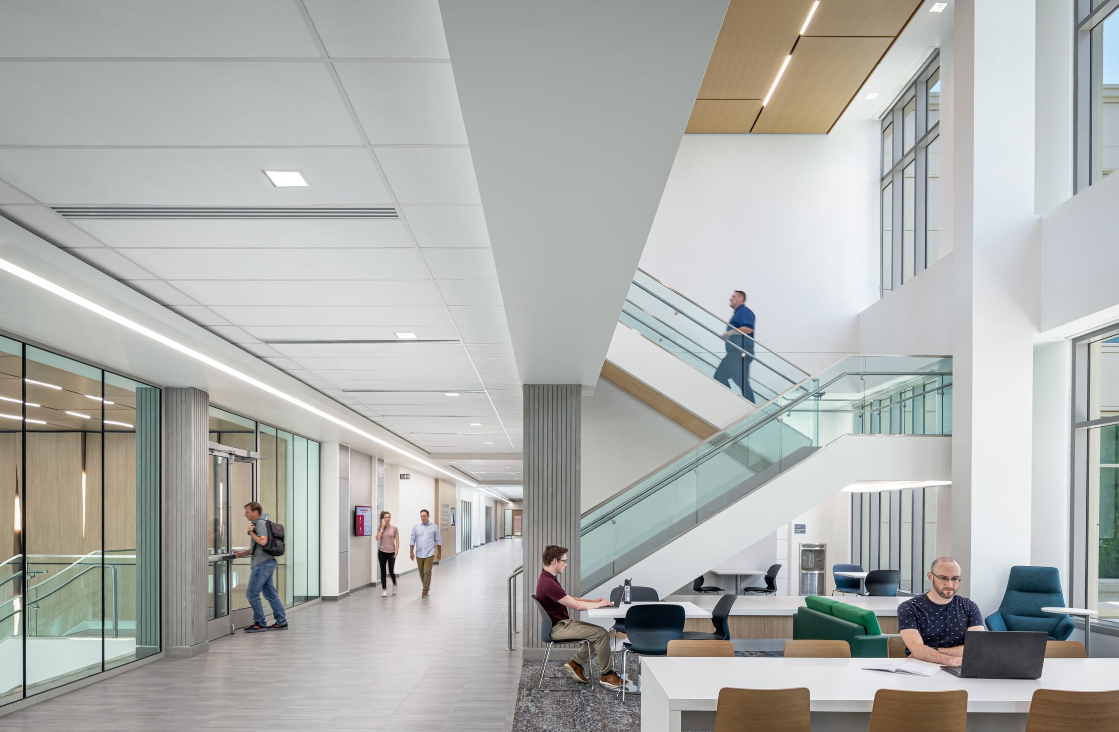 A vibrant collaborative space in the Miami University Health Sciences Building featuring a two-story atrium with modern design elements. The area includes multiple seating arrangements with wooden, black plastic, and blue fabric chairs arranged around white tables where two students work independently. A central white staircase with glass railings connects to upper levels where people are visible in motion.