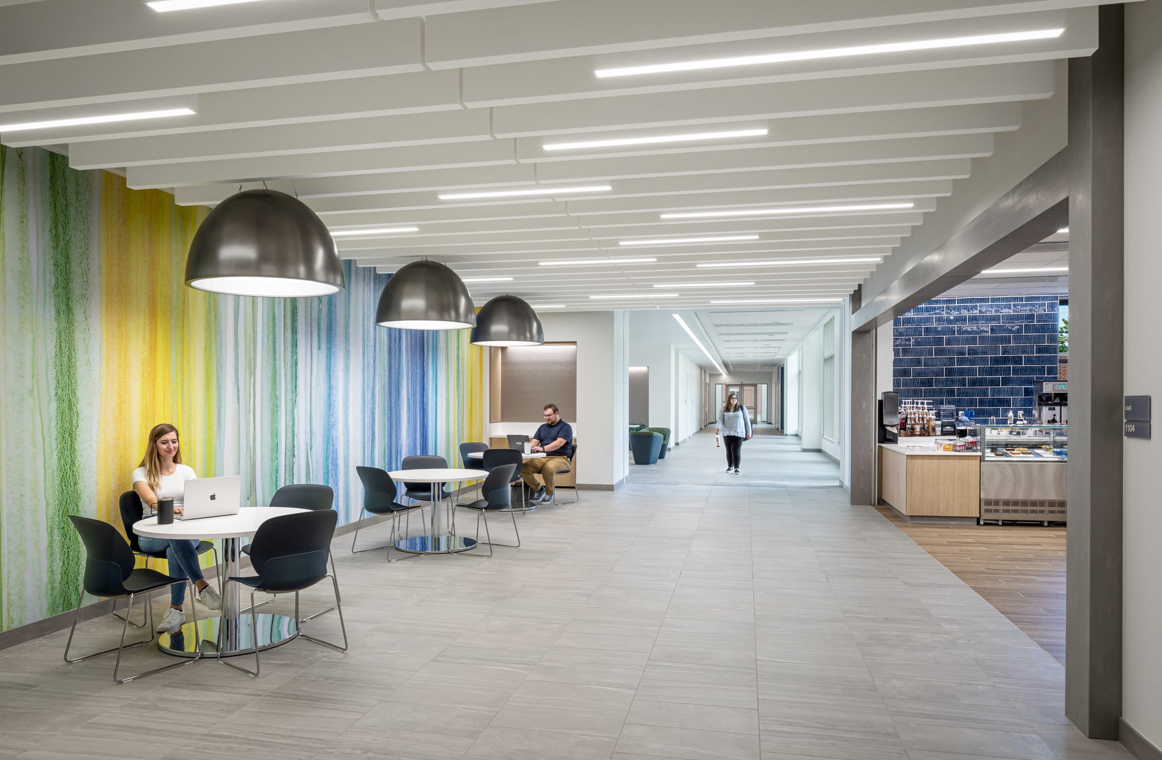 A modern café area in the Miami University Health Sciences Building featuring a vibrant watercolor-inspired wall with vertical stripes in green, yellow, and blue tones. The space includes round white tables with black chairs where students work on laptops, with large dome pendant lights overhead. A long corridor extends to the right with light-colored tile flooring and a clean, minimalist design. In the background, a person walks down the hallway, while to the right is a small café or refreshment area with a wooden counter and blue tiled wall. The bright, open space combines functional study areas with access to refreshments in a contemporary academic setting.