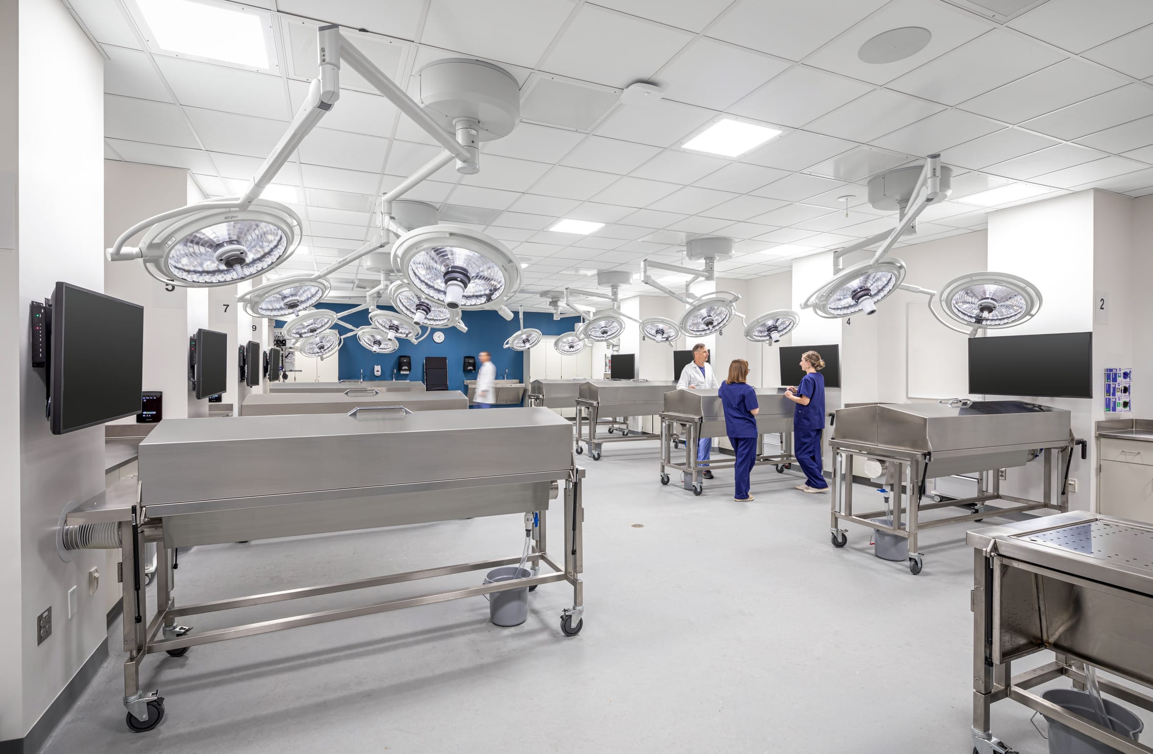 A state-of-the-art anatomy or medical simulation laboratory in Miami University's Health Sciences Building featuring multiple stainless steel examination tables with numerous ceiling-mounted surgical lights positioned above them. The clean, clinical space includes wall-mounted monitors for instruction or viewing medical imaging, and several medical professionals in blue scrubs are engaged in conversation. The room has a blue accent wall at the far end, contrasting with the predominantly white and stainless steel environment. This modern teaching facility is designed for hands-on medical education, allowing students to practice examination and procedural skills in a hospital-like setting with professional-grade equipment.