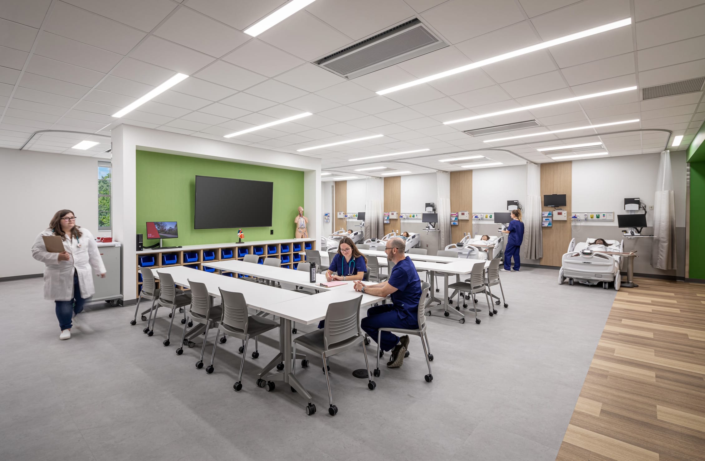 A clinical simulation training room in Miami University's Health Sciences Building that combines educational and practical medical spaces. The foreground features a classroom area with white tables and chairs where two healthcare professionals in blue scrubs are discussing notes, while another in a white coat walks nearby. A bright green accent wall displays a large monitor for instruction. The rear section contains multiple hospital bed stations with medical training mannequins, separated by privacy curtains and equipped with monitoring systems, where students practice patient care scenarios. The modern space integrates classroom learning with hands-on clinical training, allowing students to move seamlessly between theoretical discussion and practical application in a realistic healthcare environment.