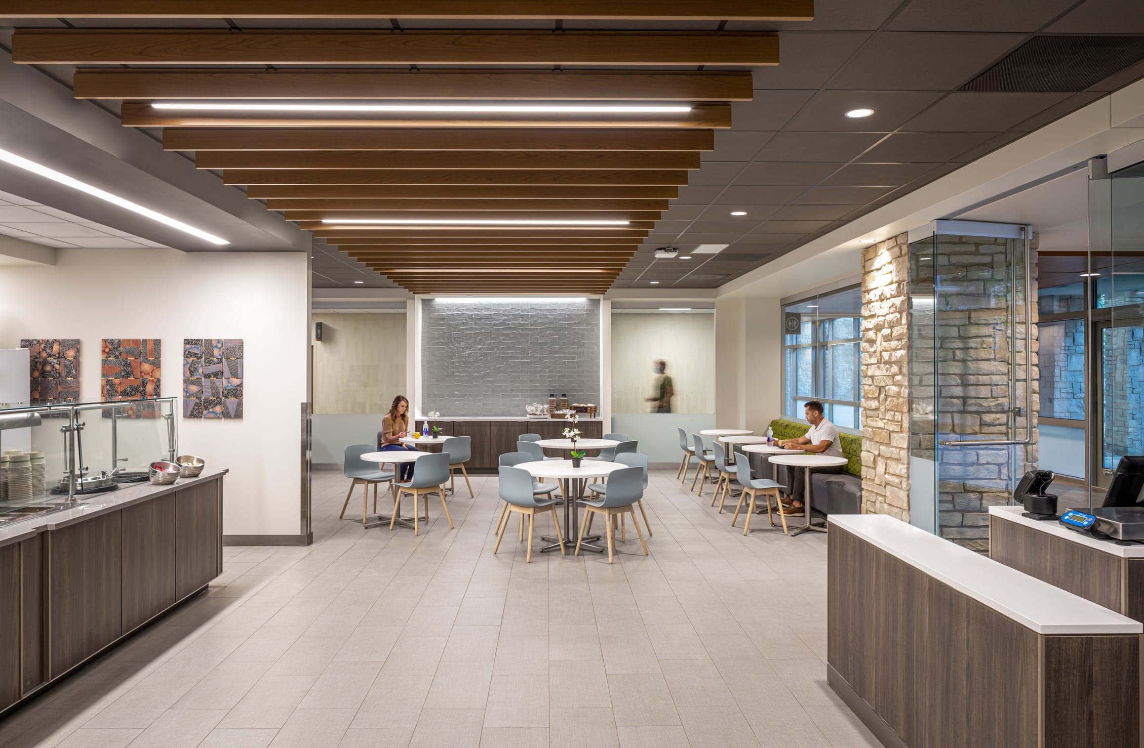 This image shows a sophisticated modern office cafeteria or break room with a sleek, contemporary design at IU Health West. The space features a dramatic ceiling treatment with parallel wood slats integrated with LED linear lighting that creates visual warmth and interest. The room includes multiple seating areas with round white tables surrounded by pale blue chairs with wooden legs. A serving counter with modern appliances and food displays runs along the left side, while a stone accent wall adds natural texture to the space. Large windows with views to the exterior bring in natural light, and colorful abstract artwork adorns the white walls. The flooring appears to be large format tiles in a neutral tone, and the overall design palette combines warm wood tones with cool grays and blues to create an inviting, healing dining environment.