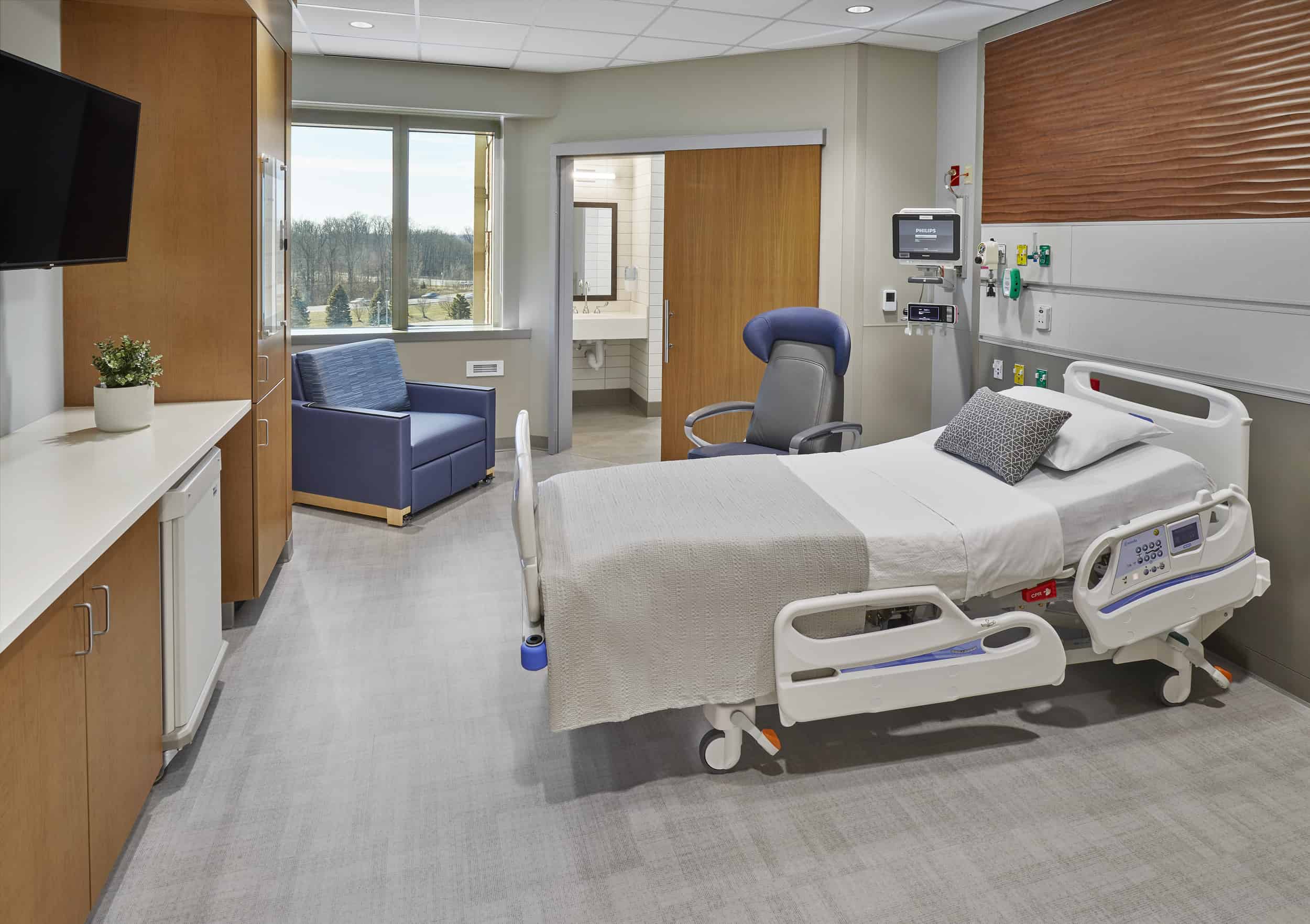 This image shows a modern, well-appointed hospital patient room designed with both functionality and comfort in mind. The room features a fully adjustable hospital bed with clean white linens and a patterned throw pillow, positioned near a large window that provides natural light and views of the landscaped grounds outside. A comfortable blue sleeper chair offers seating for visitors or family members staying overnight, while built-in wood cabinetry with a white countertop provides storage and workspace. The room includes essential medical equipment mounted on the wall behind the bed, including monitoring systems and medical gas outlets, as well as a wall-mounted television for patient entertainment. The en-suite bathroom is visible through a doorway, and the overall design uses warm wood tones, soothing blue accents, and neutral colors to create a healing environment that feels more like a hotel room than a traditional clinical setting.
