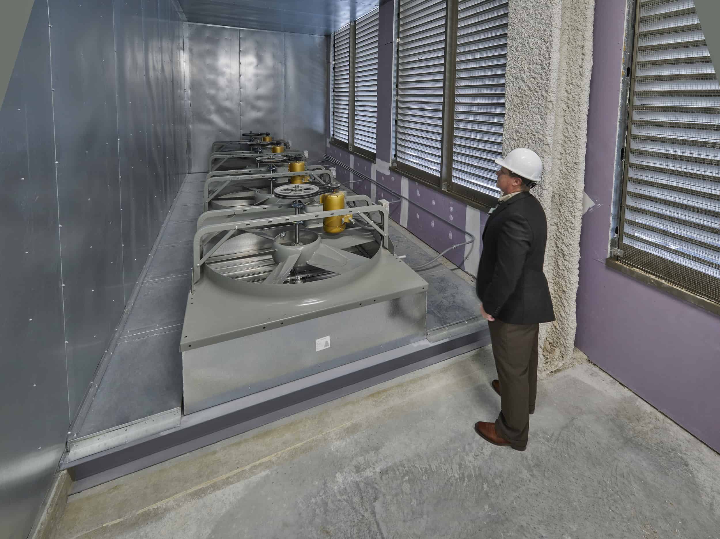 A BSA engineer in a white hard hat and dark jacket inspects a row of large industrial HVAC units housed in a mechanical room. The equipment appears to be rooftop air conditioning or ventilation units with prominent circular fans and yellow control components, all mounted on a concrete platform. The room features metal louvers along the windows for ventilation, stainless steel wall panels, and a concrete floor, creating a typical hospital building mechanical space environment.