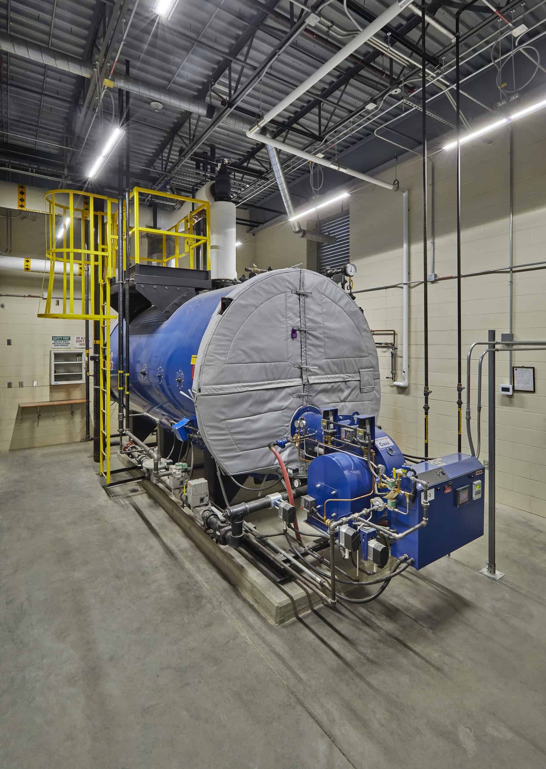 A large industrial boiler system dominates this mechanical room, featuring a cylindrical pressure vessel with gray insulation blankets and blue mechanical components. The unit is equipped with yellow safety railings and platforms for maintenance access, along with various pumps, valves, and control systems mounted on a concrete pad. The facility has an exposed ceiling structure with industrial lighting, concrete block walls, and is part of the IU Health West Hospital building's heating plant.