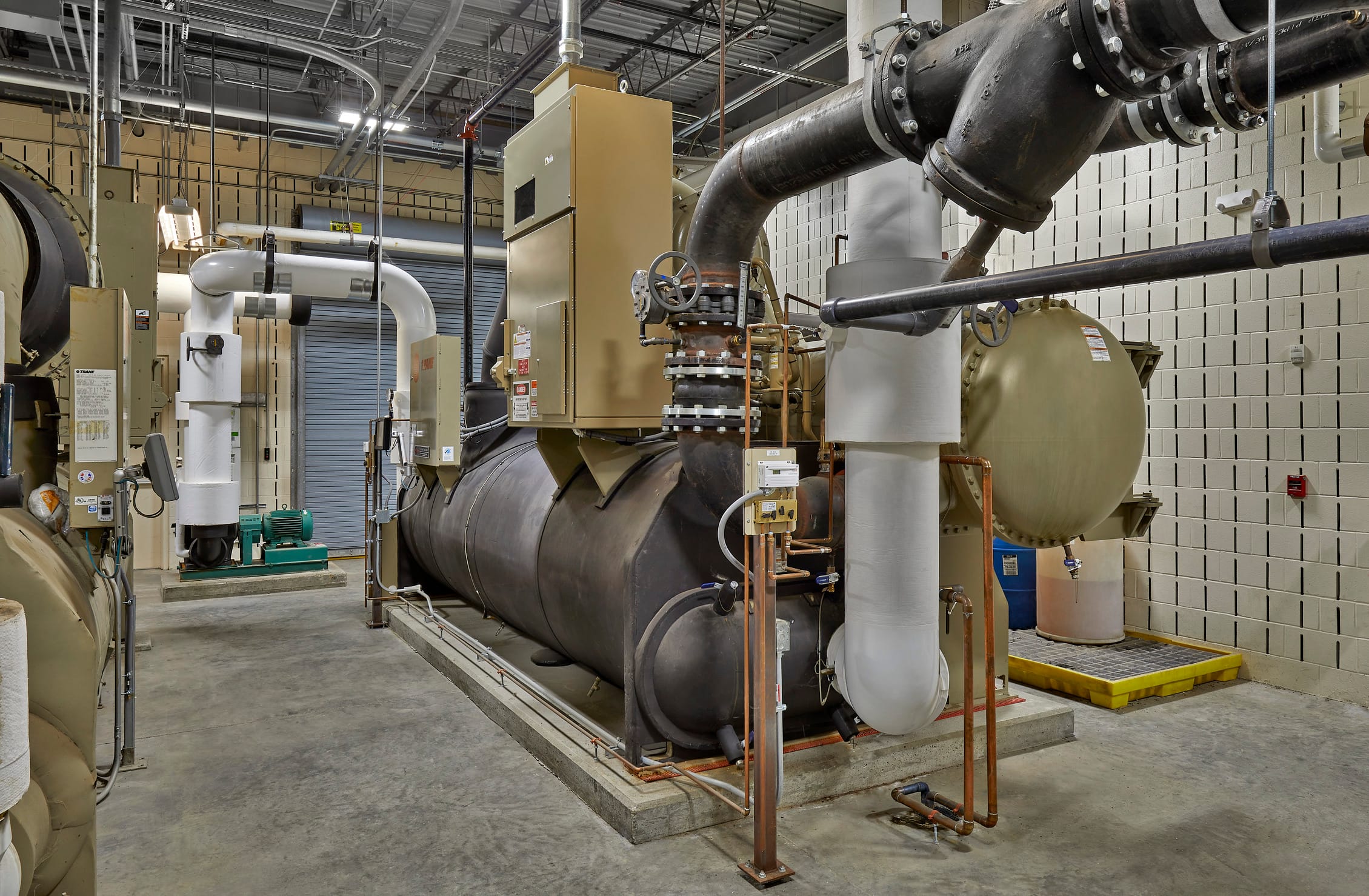 This image shows a sophisticated mechanical equipment room or boiler plant featuring large-scale HVAC infrastructure. The space contains multiple industrial boilers, chillers, and heating equipment with extensive piping systems, including both insulated and uninsulated pipes running throughout the ceiling and connecting various components. The equipment appears to be a mix of newer and older units, with beige and gray color schemes, and includes various gauges, control panels, and safety equipment. The room has an exposed ceiling with visible ductwork and lighting fixtures, concrete flooring, and white tile walls that provide a clean, functional environment for maintenance access. This type of mechanical room is designed specifically for IU Health West, where centralized heating, cooling, and hot water systems are essential for building operations.