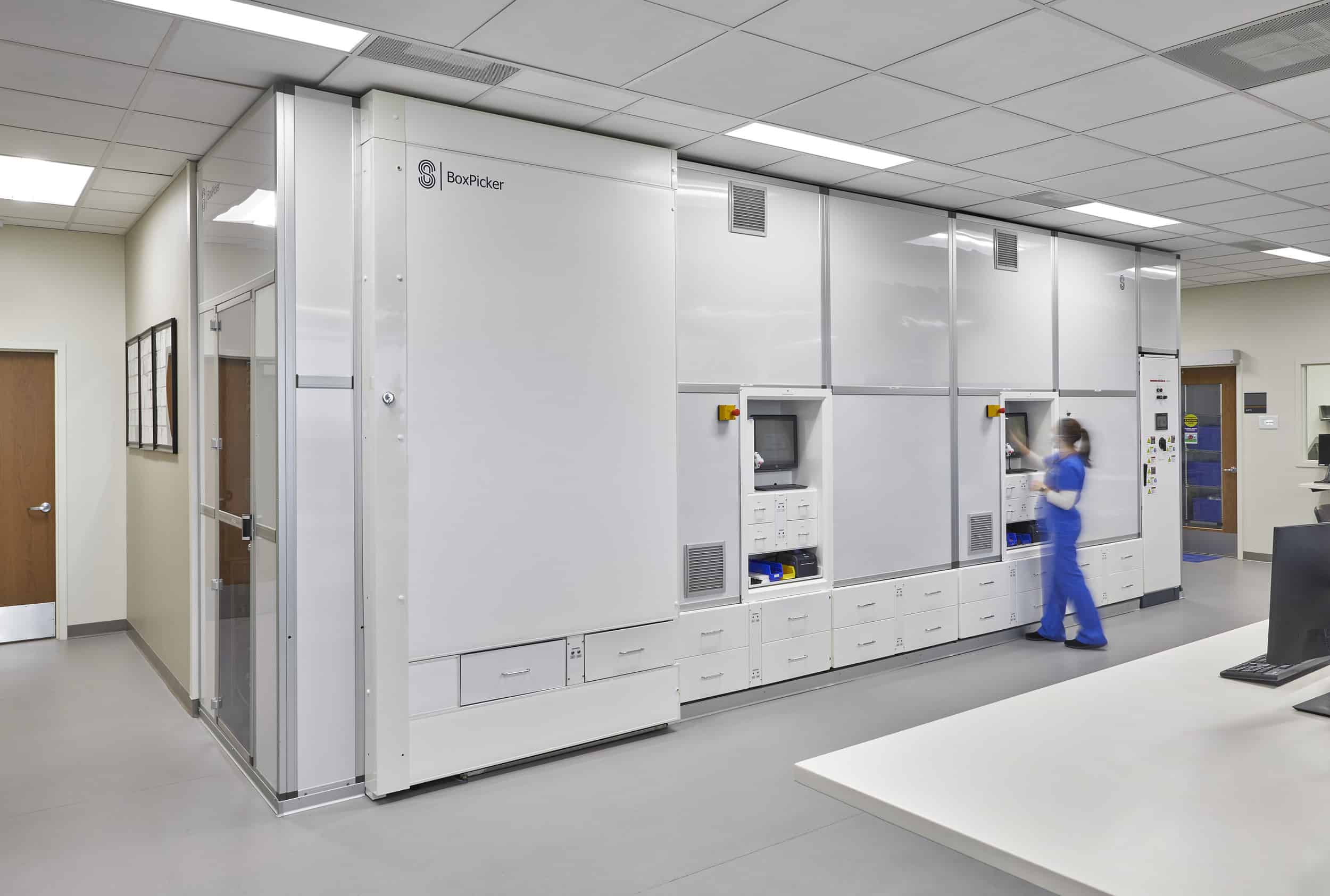 An IU Health healthcare worker in blue scrubs operates a large automated pharmacy dispensing system called 