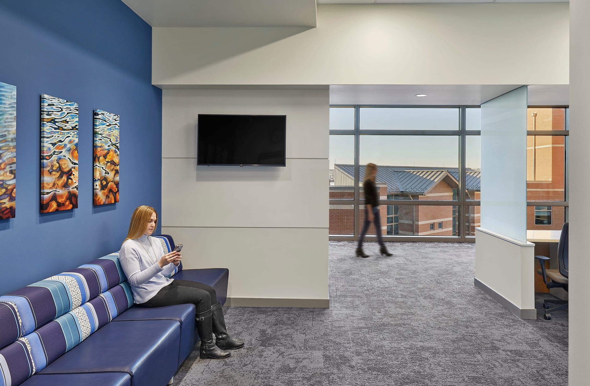 This image depicts a modern waiting area at IU Health West with a contemporary design featuring a striking blue accent wall that displays colorful abstract artwork with flowing, organic patterns in warm tones of orange and yellow. A woman sits comfortably on a sectional sofa upholstered in coordinating blue tones with geometric striped patterns, using her mobile device. The space includes a wall-mounted flat-screen TV and large floor-to-ceiling windows that provide views of brick buildings outside and allow natural light to fill the area.