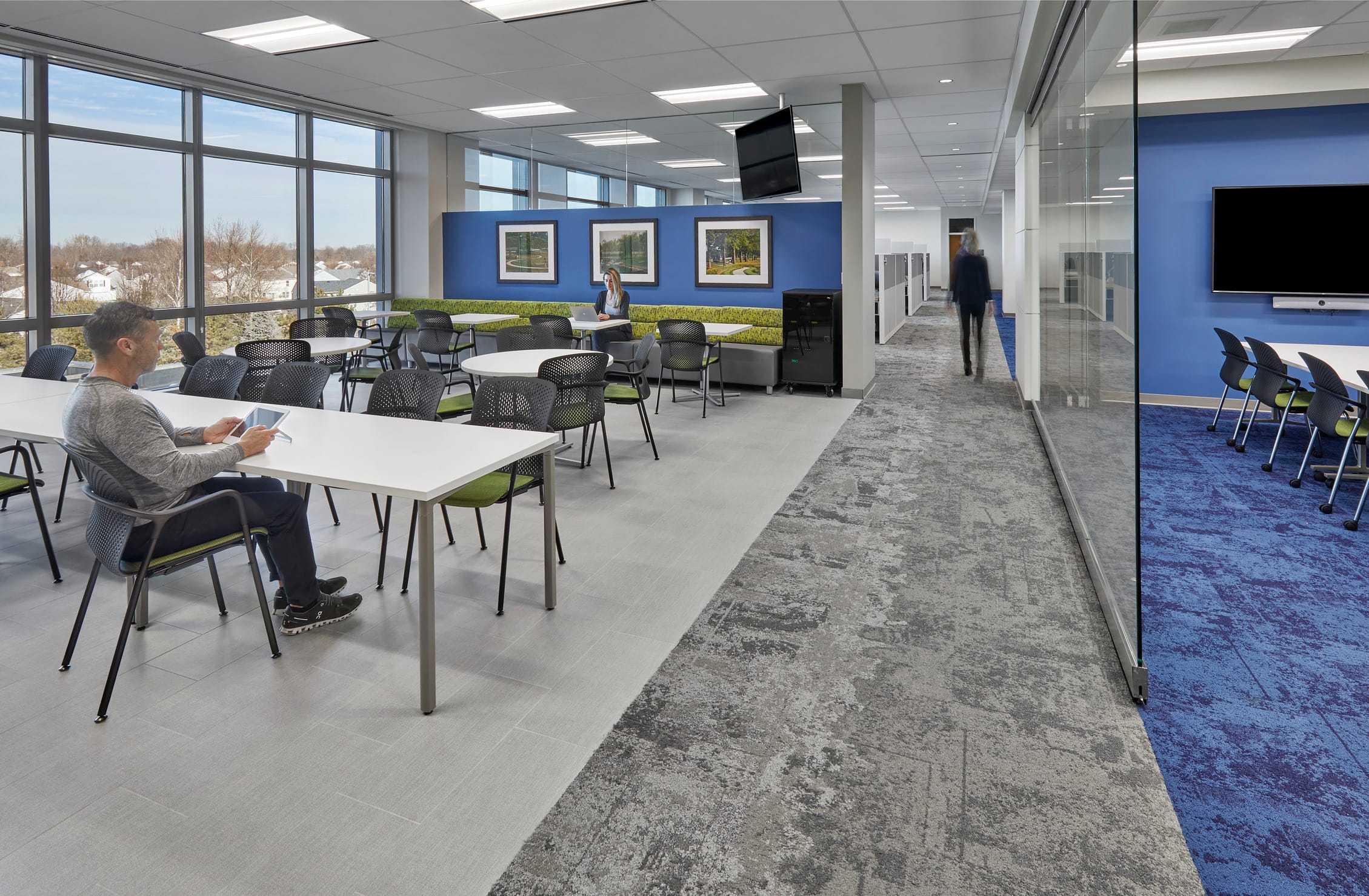 This image shows a modern multi-purpose space that combines a casual work area with adjacent conference facilities at IU Health West. The main area features white tables with black mesh chairs where people can work individually or in small groups, including a man seated at a table using a tablet. A striking blue accent wall displays three framed landscape photographs and includes built-in bench seating with green upholstery for informal meetings or relaxation. Large floor-to-ceiling windows provide abundant natural light and views of the surrounding residential neighborhood. The space flows seamlessly into a glass-enclosed conference room with blue carpeting and wall-mounted displays, while gray patterned carpet in the circulation areas helps define different zones. The design creates a flexible environment that supports various work styles, from focused individual tasks to collaborative meetings, all within a bright, contemporary setting that promotes both productivity and comfort.