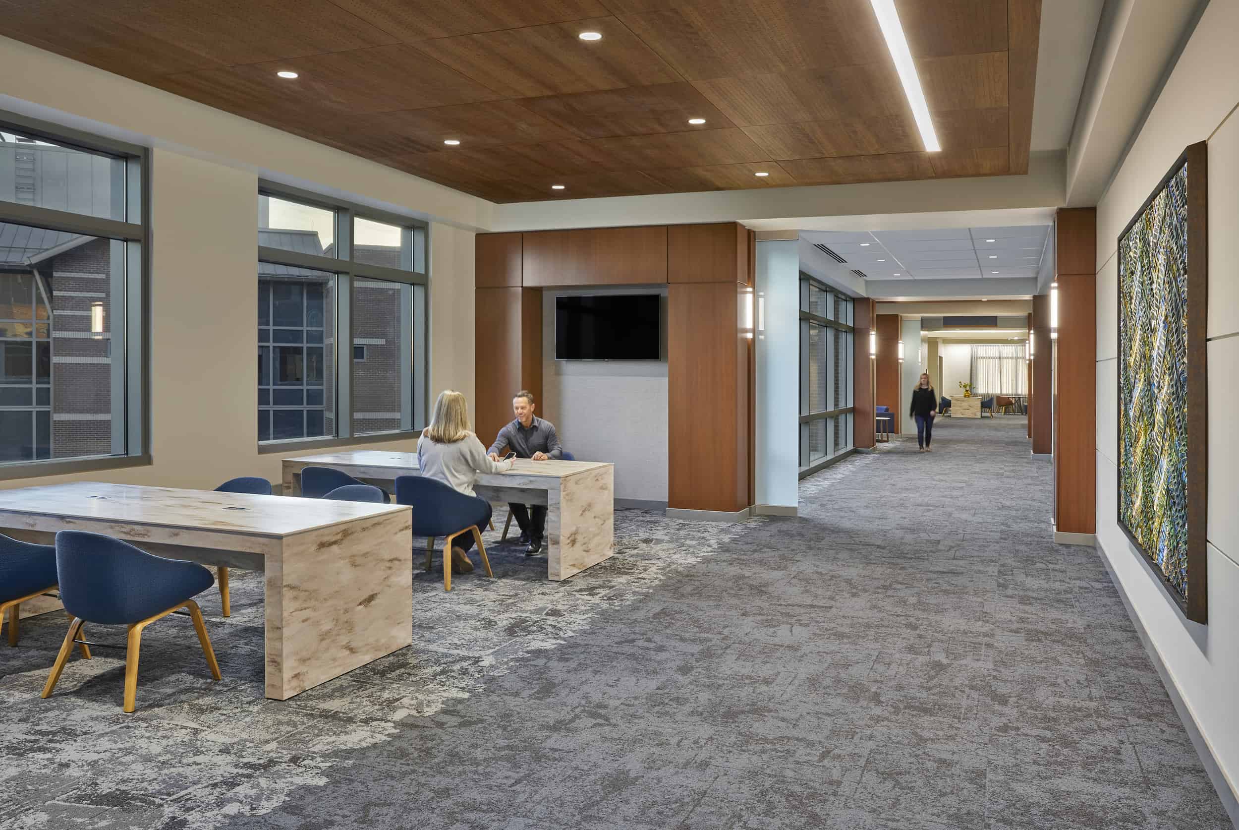 Two people meet at a modern marble table with navy blue chairs in the contemporary lobby of IU Health West Hospital, featuring a striking wood-paneled ceiling with recessed lighting, large windows overlooking adjacent buildings, and warm wood accents throughout the space. The open floor plan connects to additional seating areas and corridors, with decorative artwork on the walls and polished stone flooring creating an welcoming healthcare environment.