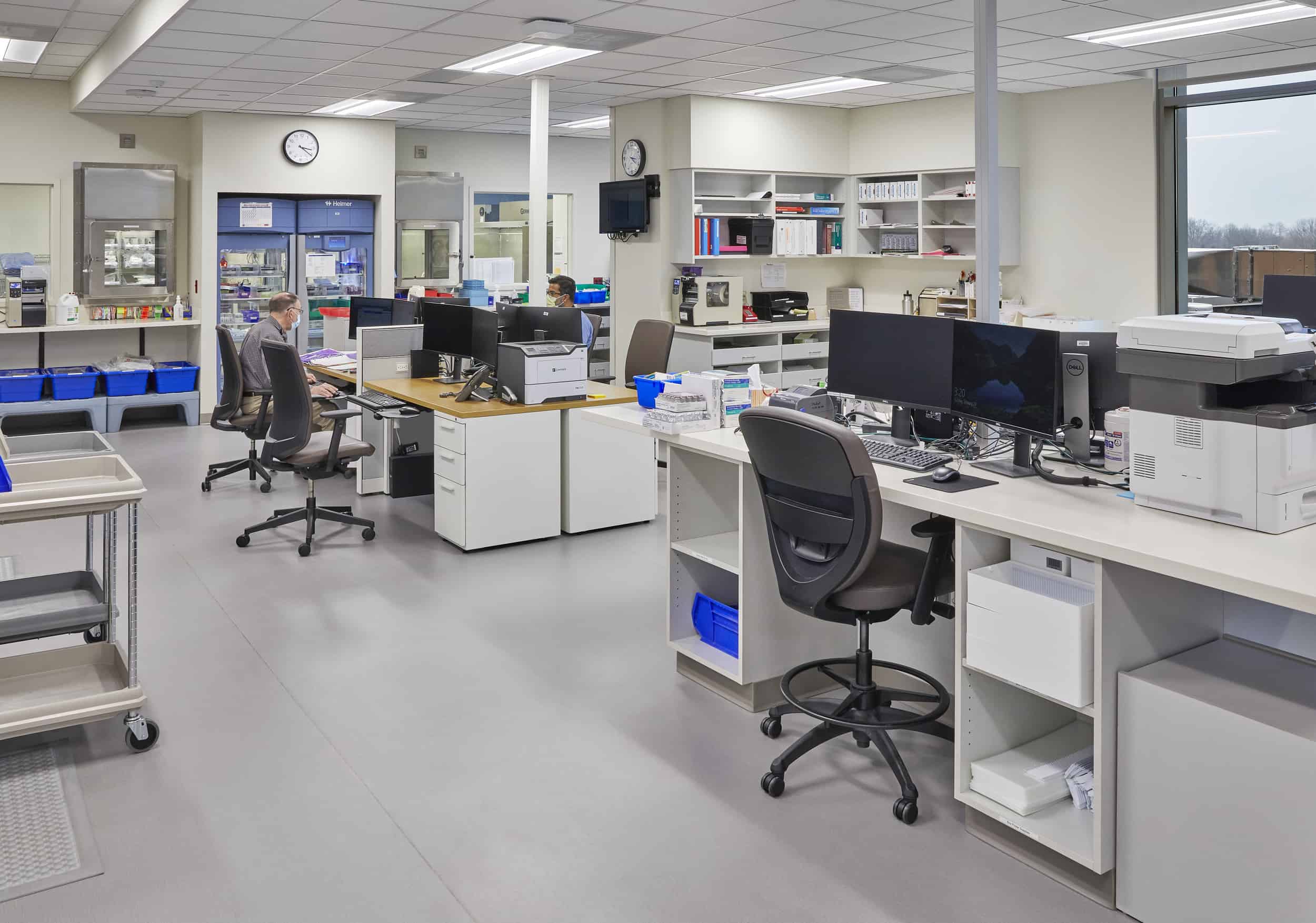 A modern hospital laboratory at IU Health West Hospital features multiple workstations with computer terminals, laboratory equipment, and storage systems arranged in an open floor plan. The clean, white environment includes adjustable office chairs, overhead shelving for supplies and reference materials, mobile carts for equipment transport, and large windows providing natural light, while a technician works at one of the stations surrounded by the specialized equipment needed for medical testing and analysis.