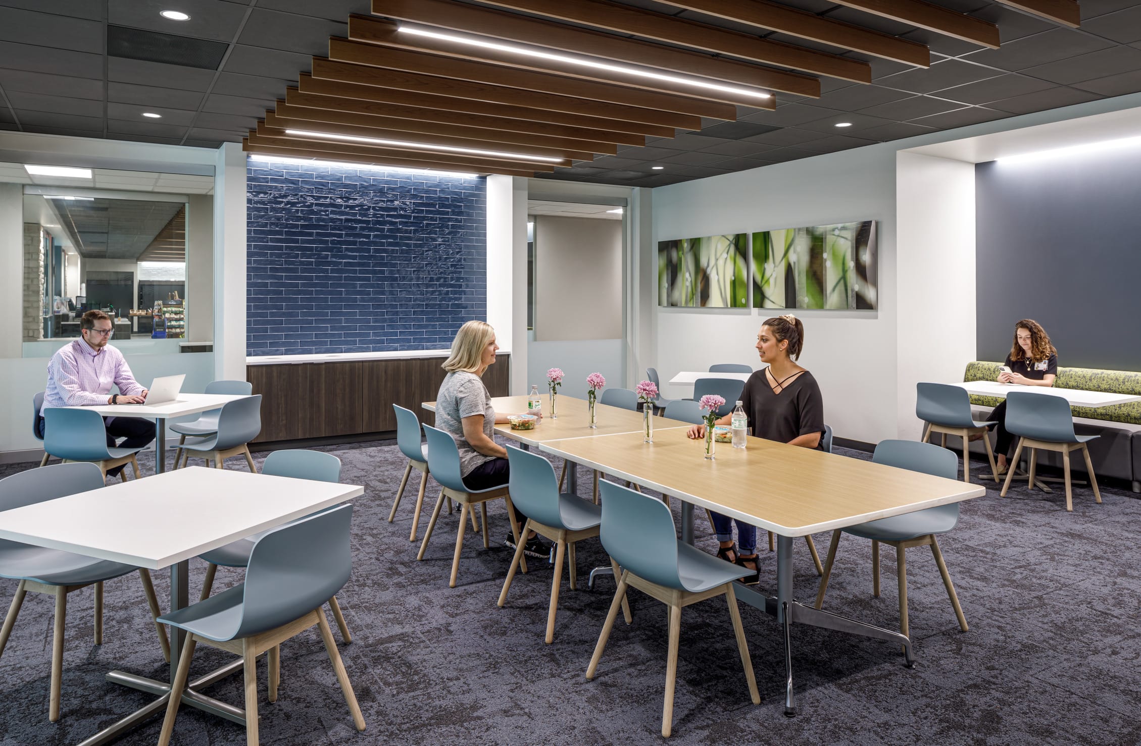 This image shows a modern cafeteria space or break room at IU Health West. The space features multiple white and light wood tables surrounded by pale blue chairs with wooden legs, where several people are seated working on laptops or having conversations. The ceiling incorporates striking linear wood slats with integrated LED lighting that creates visual interest and warmth. A navy blue subway tile accent wall serves as a dramatic backdrop, complemented by white walls displaying nature-themed artwork featuring green grasses or reeds. The flooring appears to be a textured carpet in neutral tones, and the overall color palette combines cool blues and grays with warm wood accents.