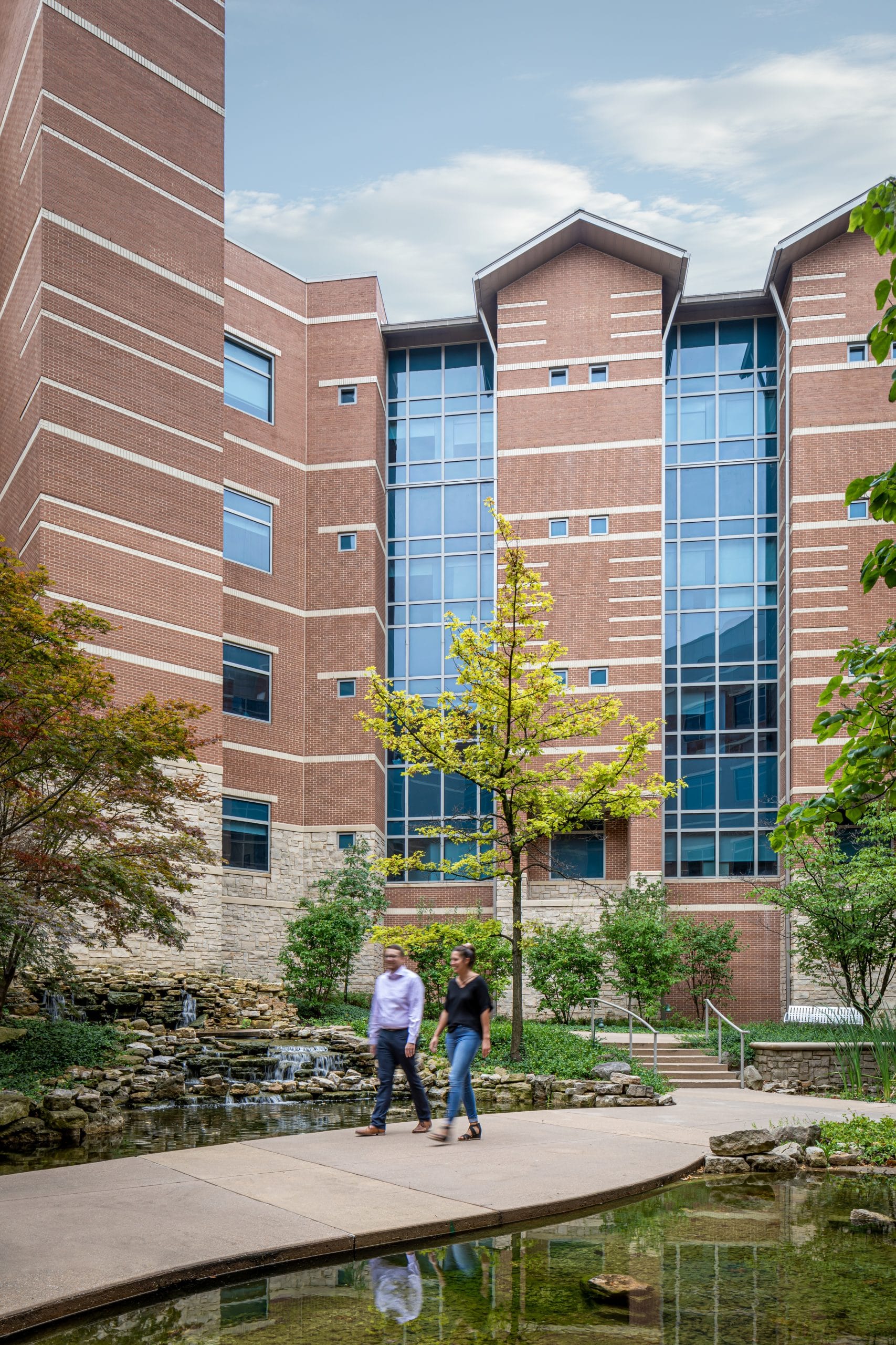 This image showcases the exterior of a modern healthcare facility, featuring a multi-story brick building with distinctive architectural elements including prominent glass curtain wall sections and white limestone accent bands. The building combines traditional red brick construction with contemporary design features, creating a professional and welcoming appearance. In the foreground, a beautifully landscaped courtyard area includes a tranquil water feature with natural stone elements, a small waterfall, and reflective pools that create a peaceful healing environment. Two people are walking along the concrete pathway that winds through the landscaped area, which is enhanced with mature trees and lush plantings. The outdoor space serves as a therapeutic garden, providing patients, visitors, and staff with a serene place for reflection and respite, while the building's design balances institutional functionality with human-scale comfort and natural beauty.