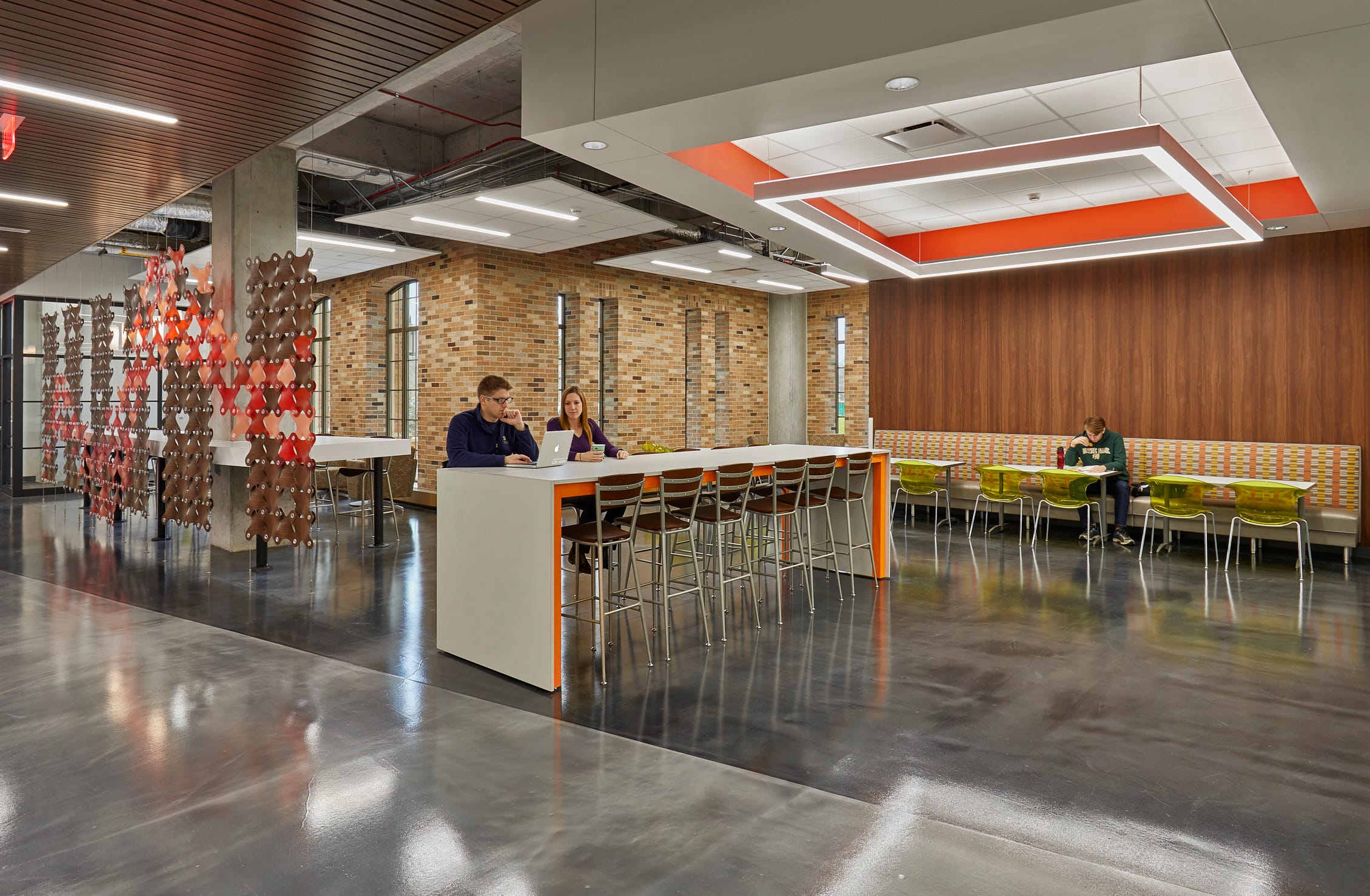 This image shows a modern university communal space with contemporary design elements. The area features a high counter workspace where two people are seated with a laptop, engaged in conversation or collaboration. The counter has bright orange accents that coordinate with the ceiling's orange rectangular lighting feature. To the right are booth-style seating areas with colorful patterned upholstery and lime green chairs, where another person can be seen working. The space has a mix of materials including polished concrete floors, exposed brick walls, and warm wood paneling on the right wall. On the left side, there's a decorative partition or screen with a geometric diamond pattern in shades of red and brown that provides partial separation between areas while maintaining an open feel. The ceiling combines wood slat sections with exposed mechanical systems, creating an industrial yet warm aesthetic. The overall design balances openness with defined zones for different activities, typical of contemporary academic buildings designed to encourage both collaboration and individual work in a casual, appealing environment.