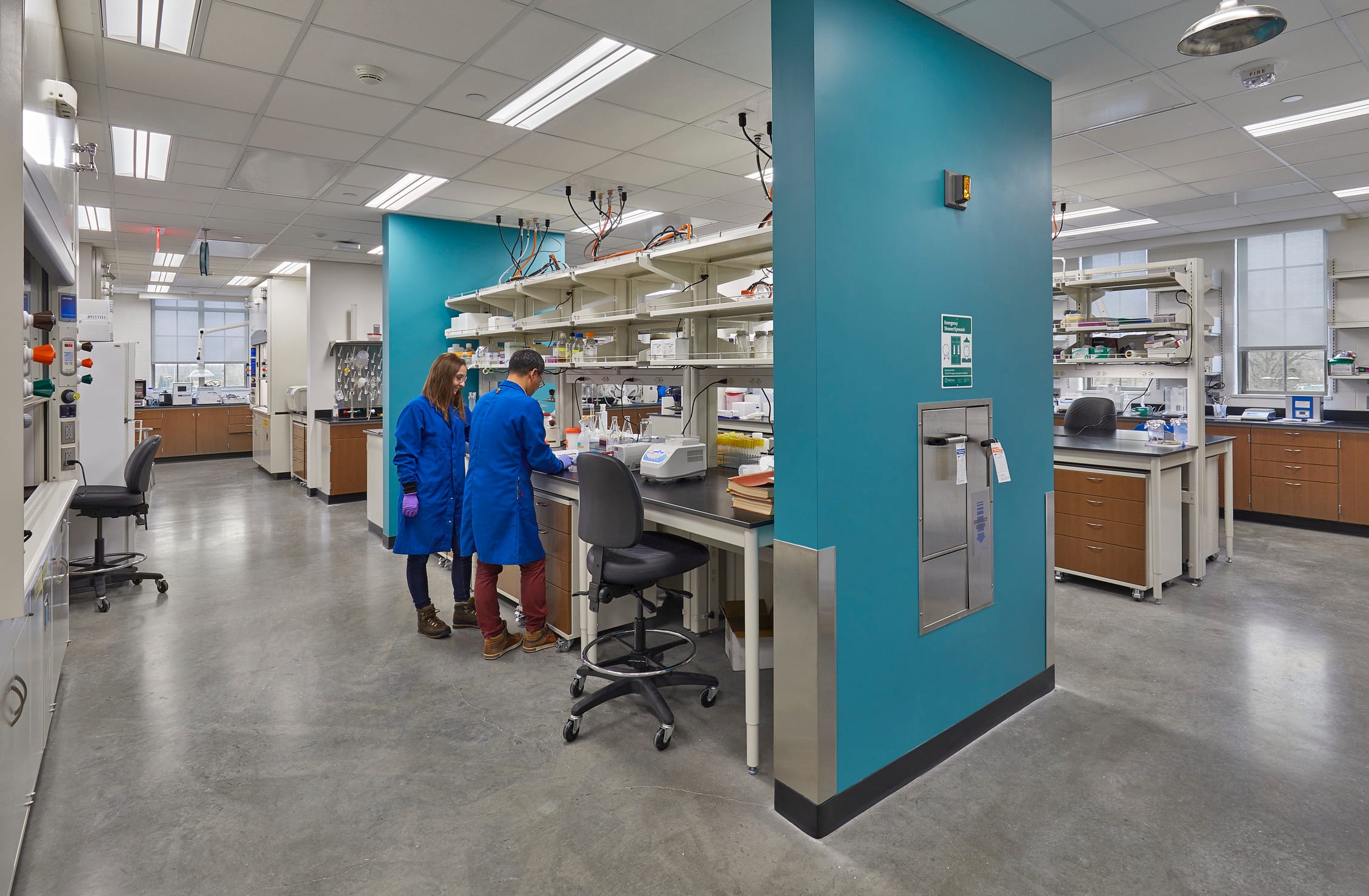The image shows a clinical laboratory. The room has a central work area with multiple shelves holding chemical containers and other lab equipment. This central workstation has teal walls at each end, forming dividers from the rest of the room. Similar workstations are in the background but are open and not separated by walls, signifying the importance of the central workstation. Two students in blue lab coats are working collaboratively near glass vials and a scale. The room has white walls, a dropped ceiling with fluorescent lighting, and polished concrete flooring.