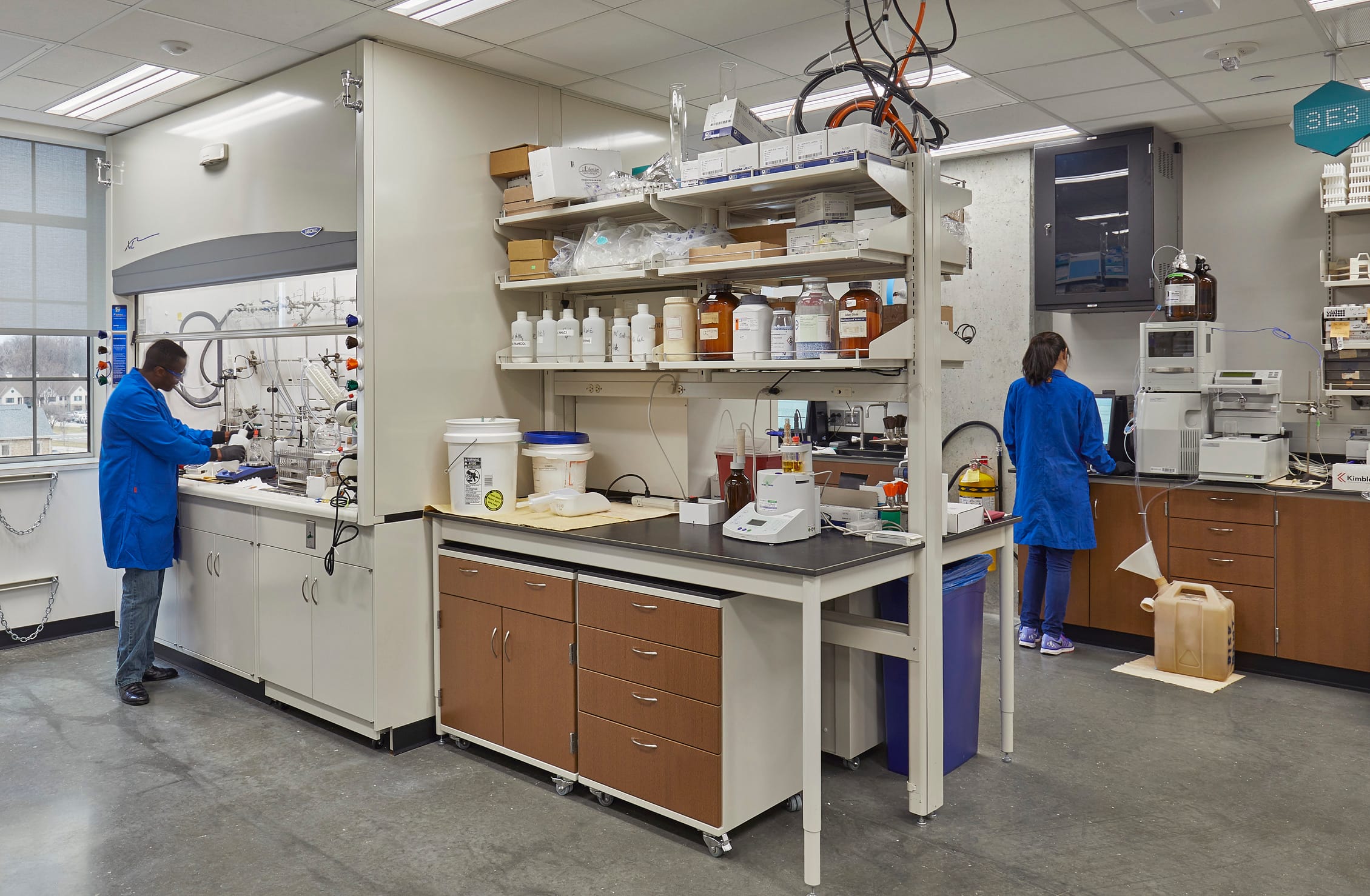 The image shows a clinical laboratory. The room has a central work area with multiple shelves holding chemical containers and other lab equipment. A student in a blue lab coat is working underneath a fume hood and pouring a liquid solution into a highly technical machine. Another student in the same style of lab coat is working on a computer. The room has white walls, a dropped ceiling with fluorescent lighting, and polished concrete flooring.