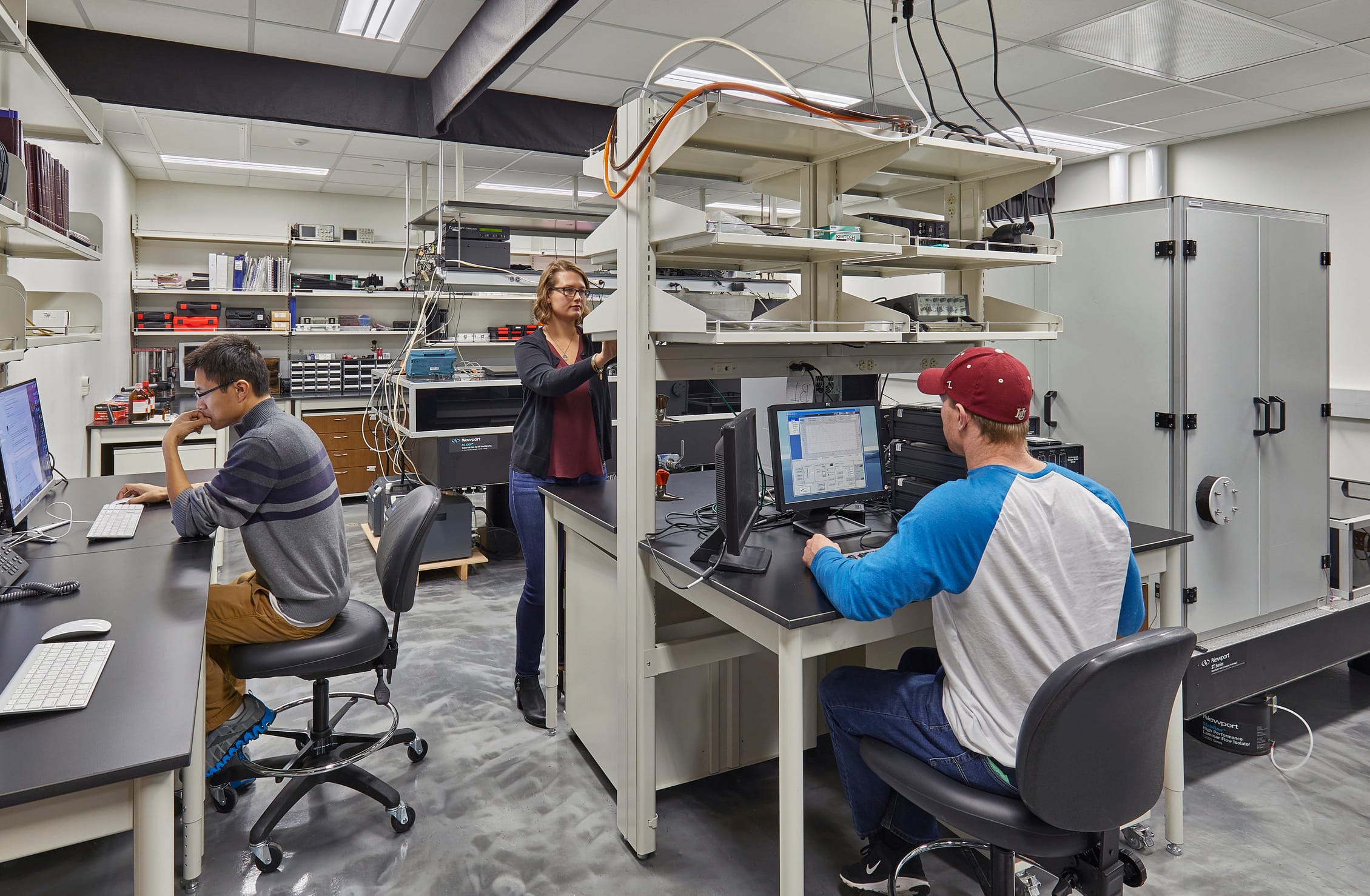 The image shows a laboratory or technical workspace with three people working at computer stations. The room has a central equipment rack or workbench with multiple shelves containing technical equipment and wiring. One person with glasses wearing a gray striped shirt is seated on the left side at a computer workstation. In the center, a person with shoulder-length hair is standing and appears to be adjusting something on the central equipment rack. On the right side, another person wearing a red baseball cap and blue/white baseball-style shirt is seated at a computer workstation. The room has white walls, a dropped ceiling with fluorescent lighting, and gray flooring. The background contains shelving units with various electronic equipment, cables, and storage containers. This appears to be a technology lab.