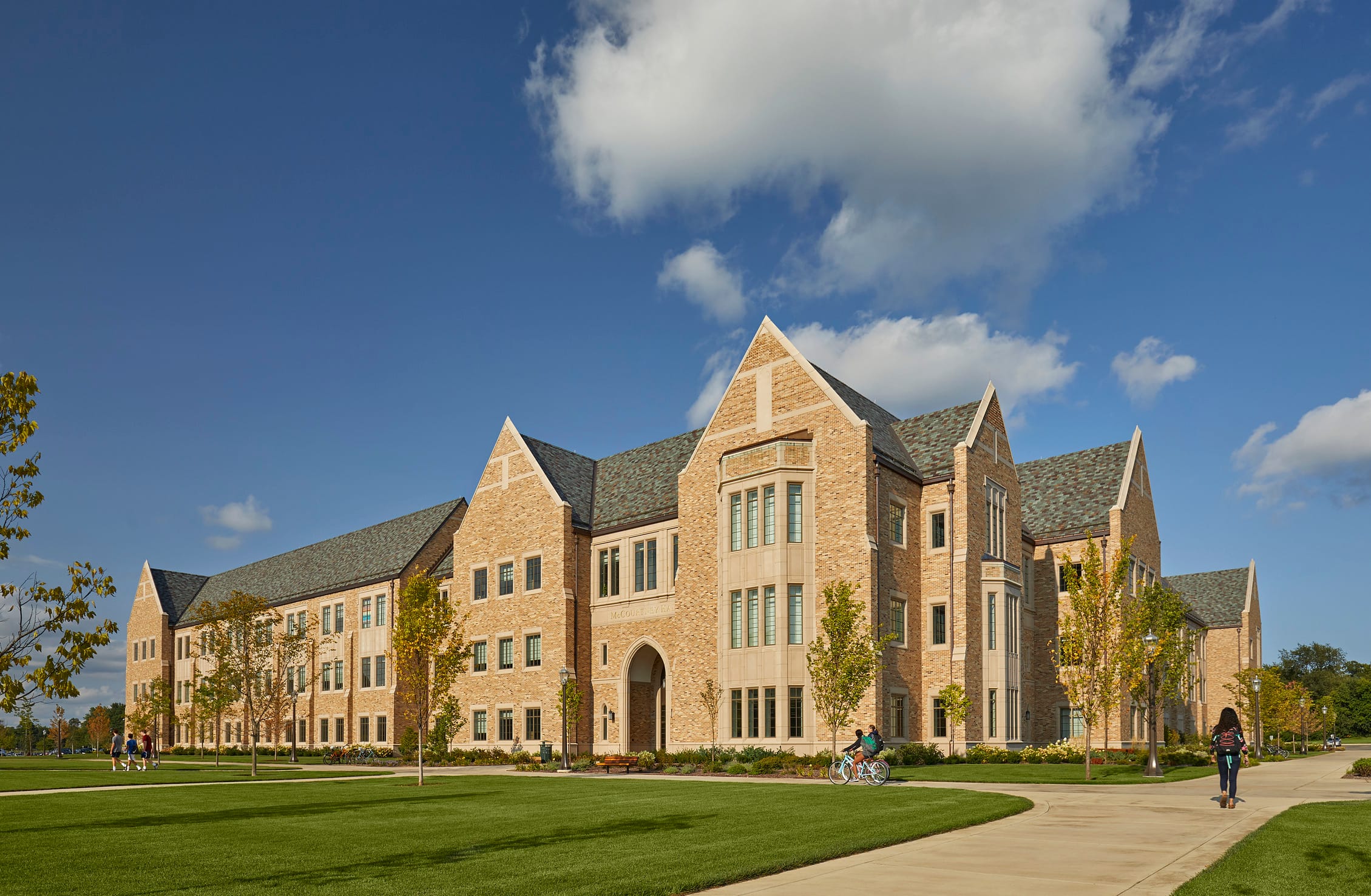 This image shows a large collegiate Gothic-style academic building on Notre Dame's campus. The structure features tan/beige brick construction with multiple peaked gables and a gray slate roof. The building is three stories tall with symmetrical wings extending from a central entrance area that appears to have an arched entryway. In the foreground, there's a well-maintained green lawn with a curved walkway. Along the walkway, students are walking and riding bikes. Young trees are planted around the perimeter of the building, suggesting relatively recent landscaping. The scene is set against a dramatic blue sky filled with white clouds. The entire scene conveys a serene, academic environment on what appears to be a pleasant day.